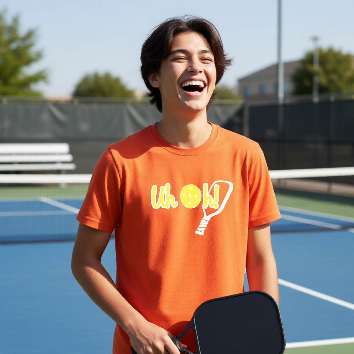A young person on a pickleball court wearing an orange short-sleeve t-shirt with a white and yellow graphic of the words “Uh Oh!” with a yellow pickleball replacing the letter O on the front of the shirt, over the top of a pickleball paddle.