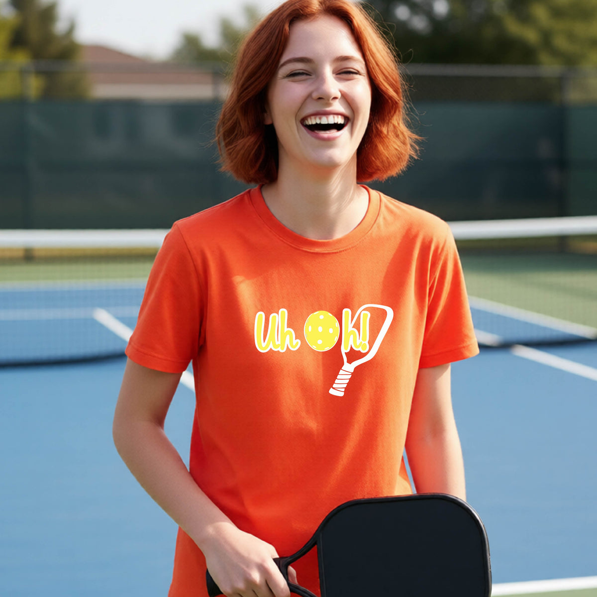 A young person on a pickleball court wearing an orange short-sleeve t-shirt with a white and yellow graphic of the words “Uh Oh!” with a yellow pickleball replacing the letter O on the front of the shirt, over the top of a pickleball paddle.
