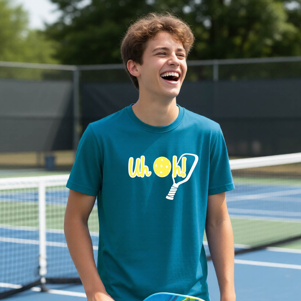 A young person on a pickleball court wearing a teal short-sleeve t-shirt with a white and yellow graphic of the words “Uh Oh!” with a yellow pickleball replacing the letter O on the front of the shirt, over the top of a pickleball paddle.