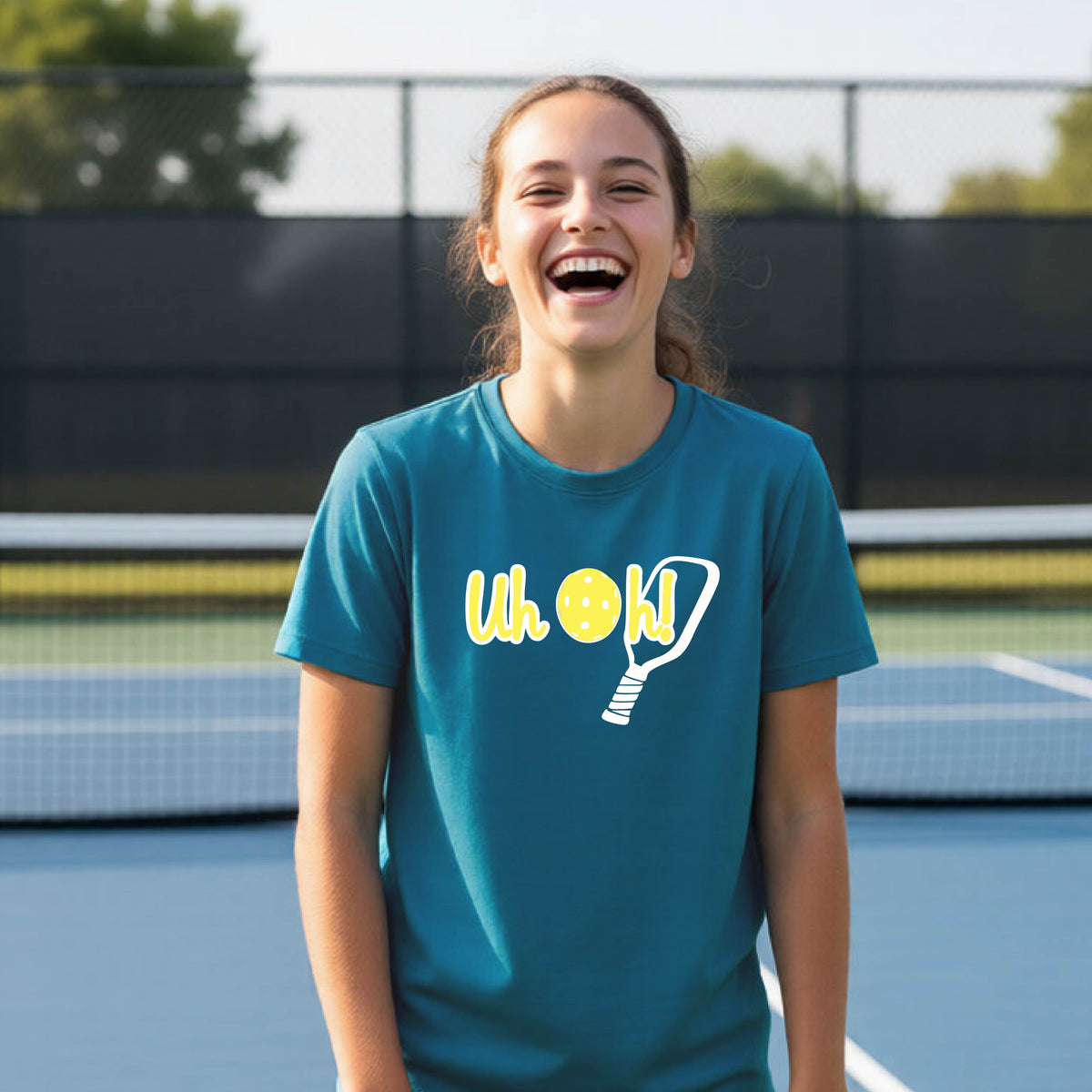 A young person on a pickleball court wearing a teal short-sleeve t-shirt with a white and yellow graphic of the words “Uh Oh!” with a yellow pickleball replacing the letter O on the front of the shirt, over the top of a pickleball paddle.