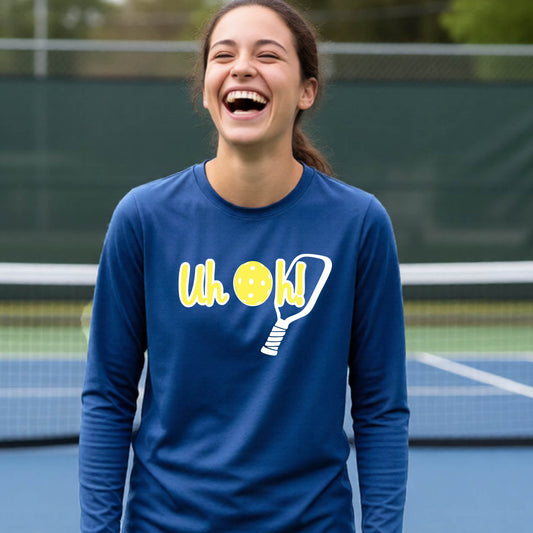 A young person on a pickleball court wearing a royal blue long-sleeve t-shirt with a white and yellow graphic of the words “Uh Oh!” with a yellow pickleball replacing the letter O on the front of the shirt, over the top of a pickleball paddle.