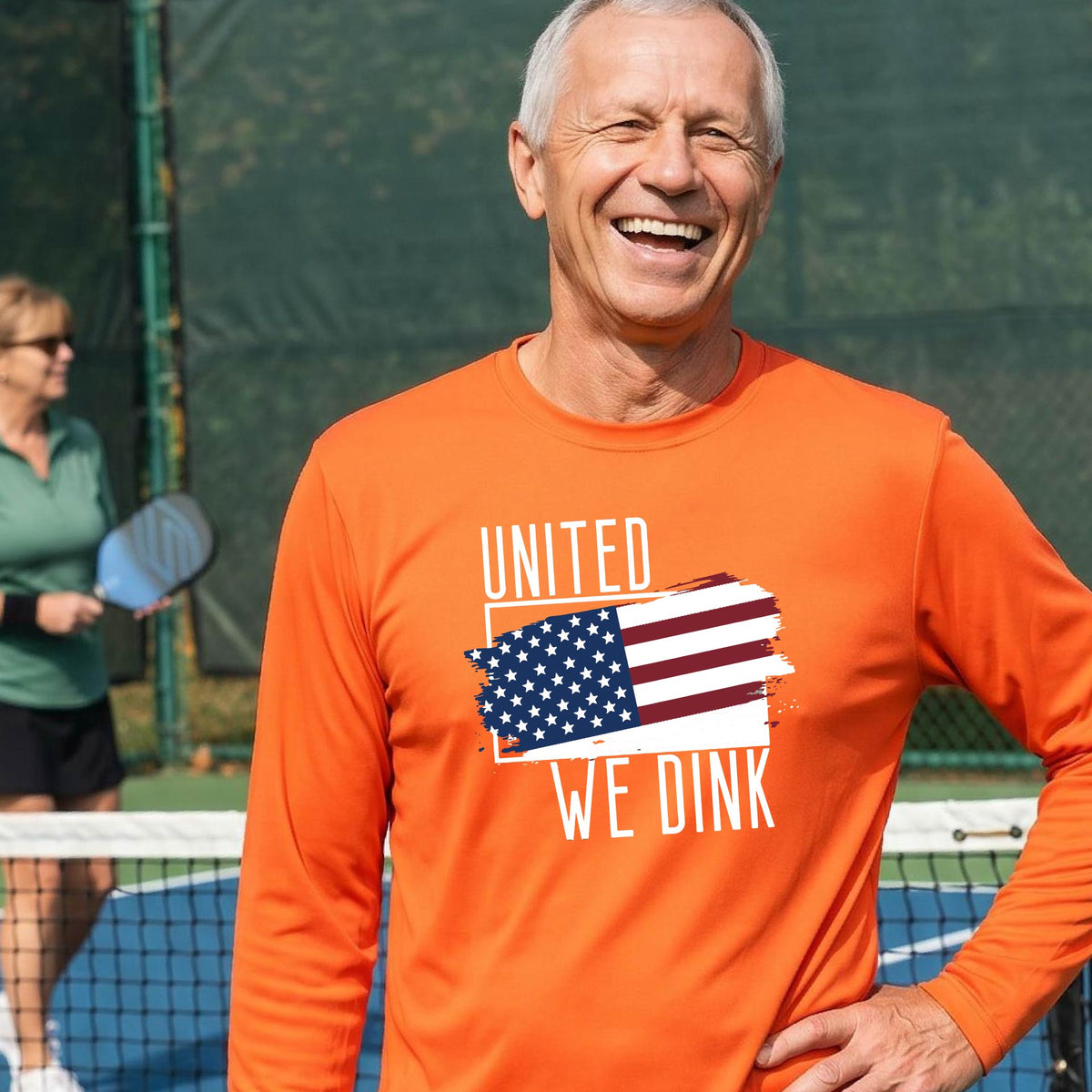 A man on a pickleball court wearing an orange long-sleeve t-shirt with a pickleball themed red, white and blue graphic of a patriotic flag with white words “UNITED WE DINK” above and below the flag on the front of the shirt.