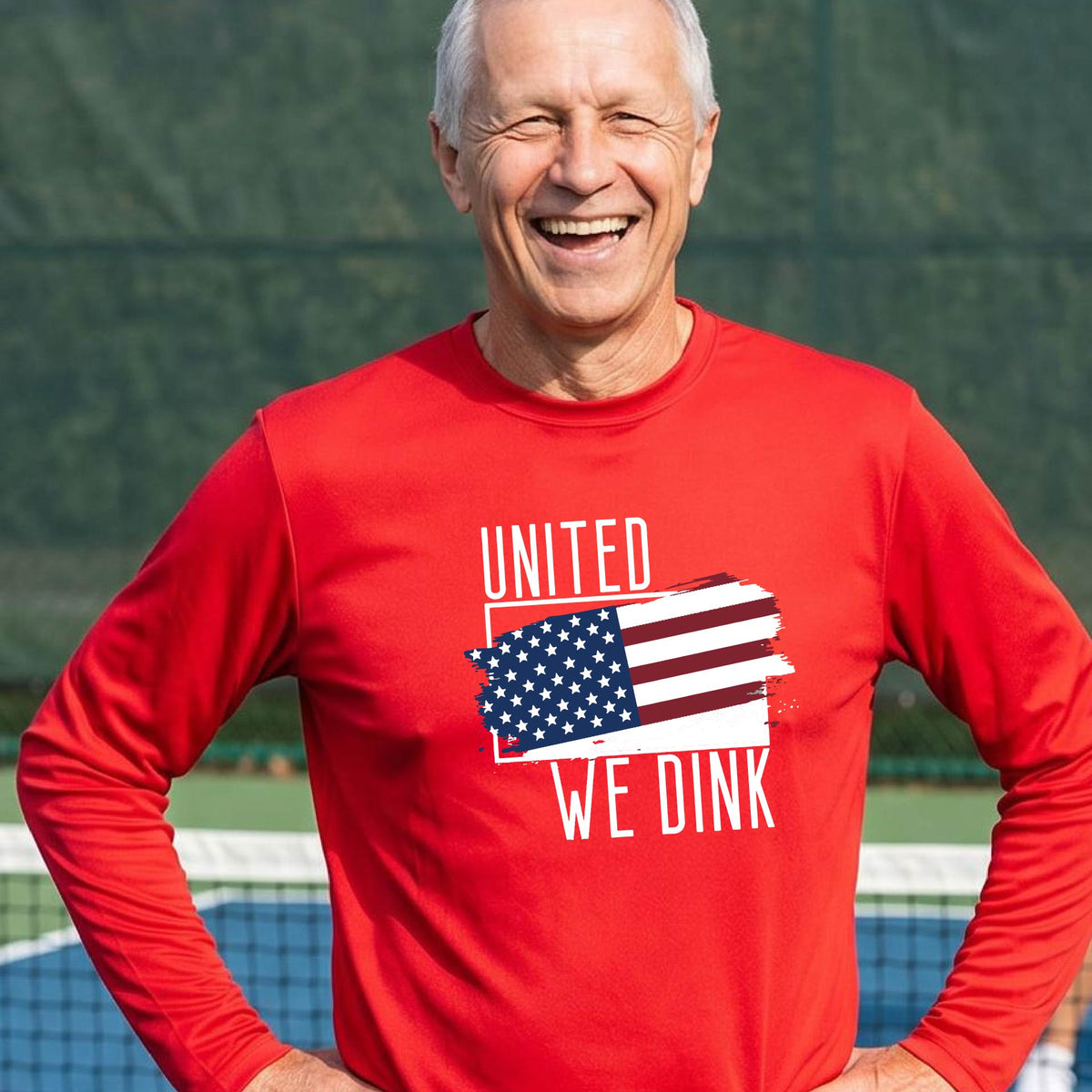 A man on a pickleball court wearing a red long-sleeve t-shirt with a pickleball themed red, white and blue graphic of a patriotic flag with white words “UNITED WE DINK” above and below the flag on the front of the shirt.