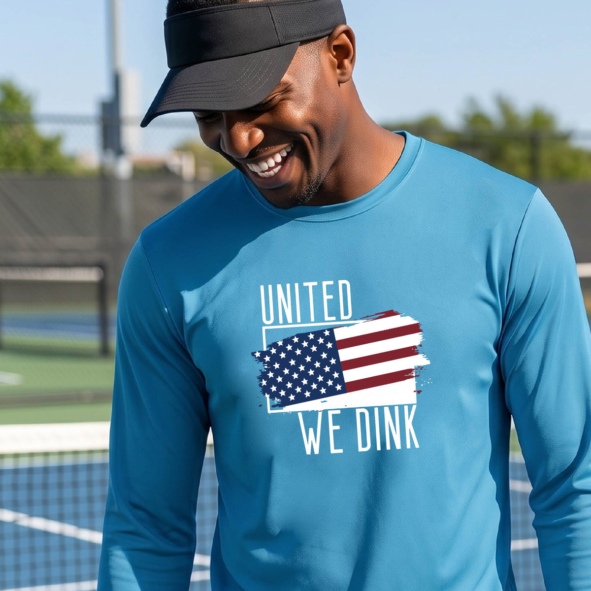 A man on a pickleball court wearing a bright blue long-sleeve t-shirt with a pickleball themed red, white and blue graphic of a patriotic flag with white words “UNITED WE DINK” above and below the flag on the front of the shirt.