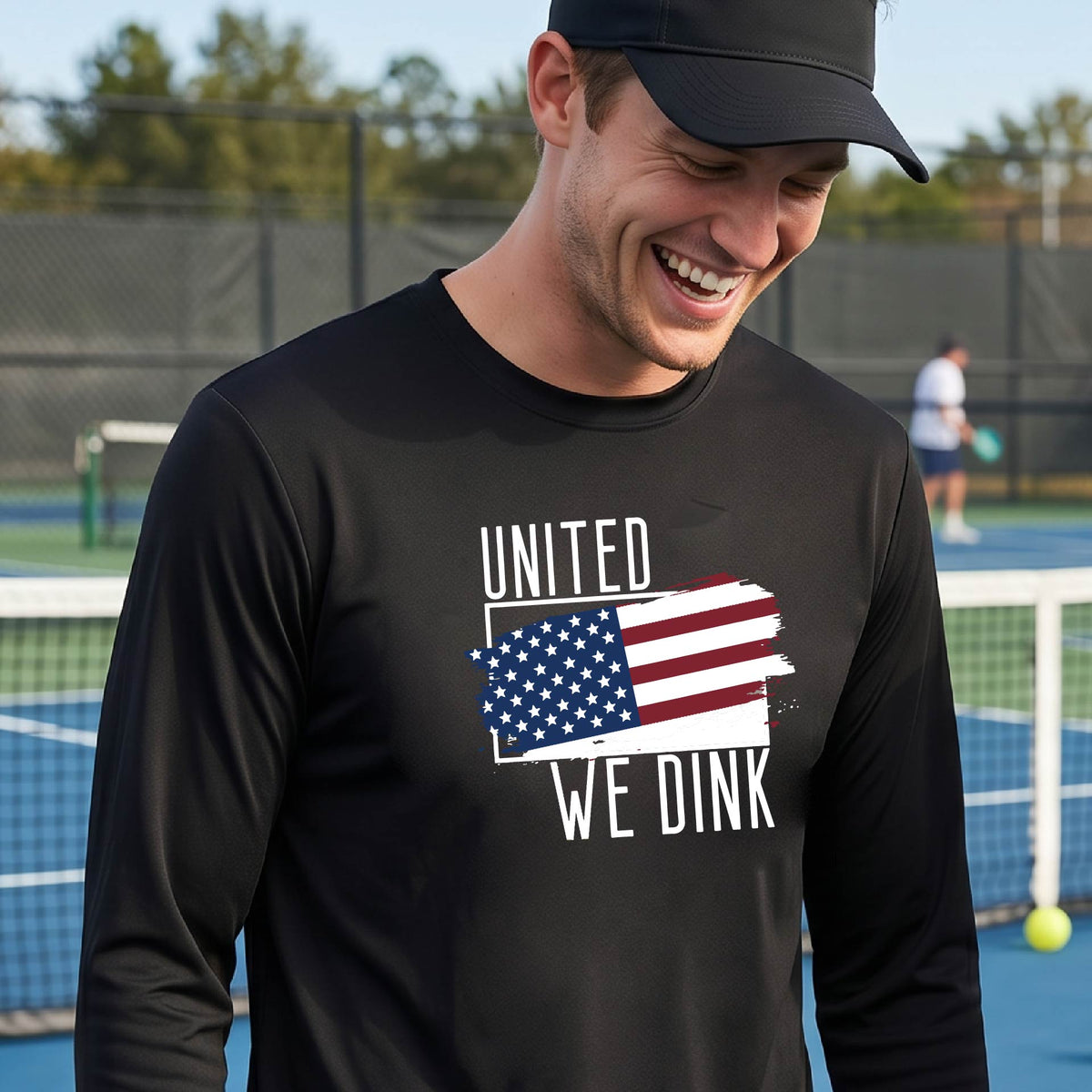 A man on a pickleball court wearing a black long-sleeve t-shirt with a pickleball themed red, white and blue graphic of a patriotic flag with white words “UNITED WE DINK” above and below the flag on the front of the shirt.