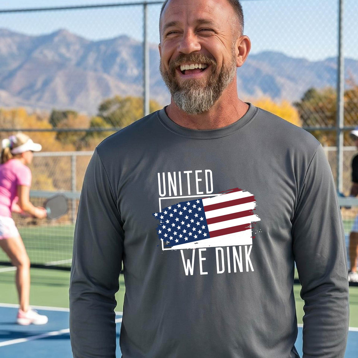 A man on a pickleball court wearing a gray long-sleeve t-shirt with a pickleball themed red, white and blue graphic of a patriotic flag with white words “UNITED WE DINK” above and below the flag on the front of the shirt.