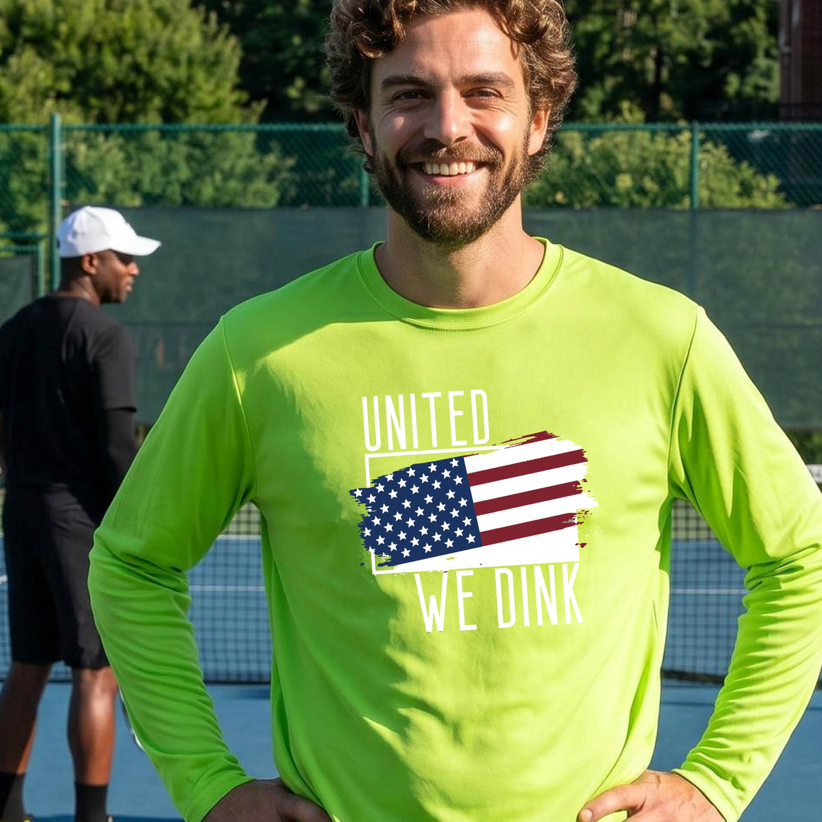 A man on a pickleball court wearing a lime green long-sleeve t-shirt with a pickleball themed red, white and blue graphic of a patriotic flag with white words “UNITED WE DINK” above and below the flag on the front of the shirt.
