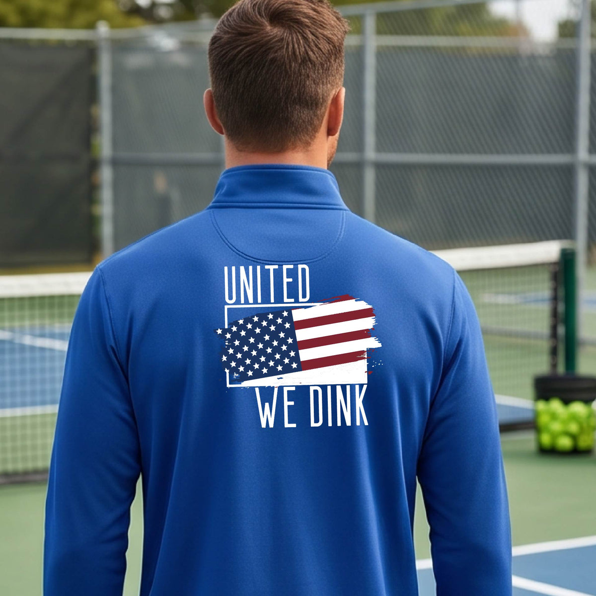 A man seen from behind on a pickleball court wearing a royal blue quarter-zip shirt with a pickleball themed red, white and blue graphic of a patriotic flag with white words “UNITED WE DINK” above and below the flag on the back of the shirt.