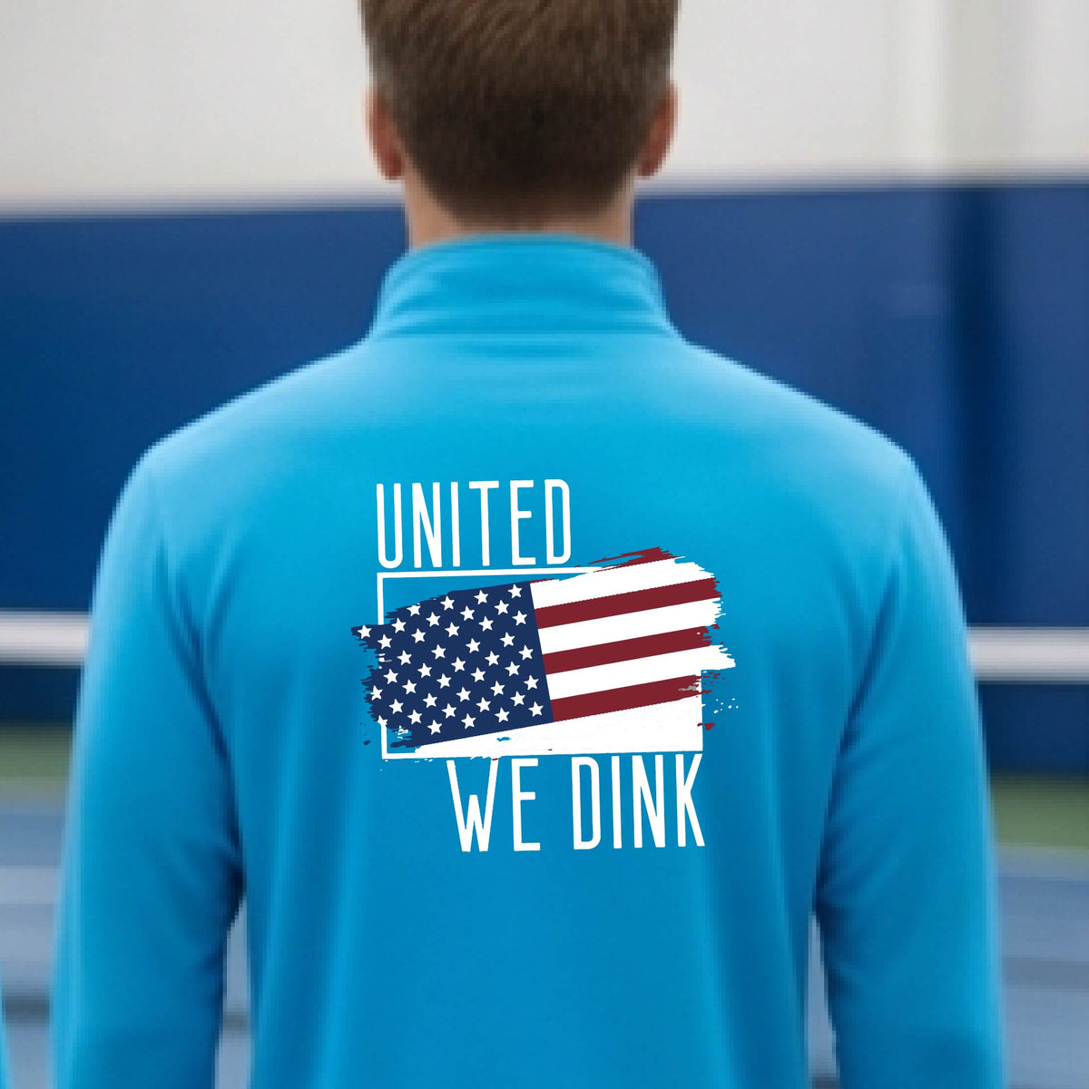 A man seen from behind on a pickleball court wearing a bright blue quarter-zip shirt with a pickleball themed red, white and blue graphic of a patriotic flag with white words “UNITED WE DINK” above and below the flag on the back of the shirt.