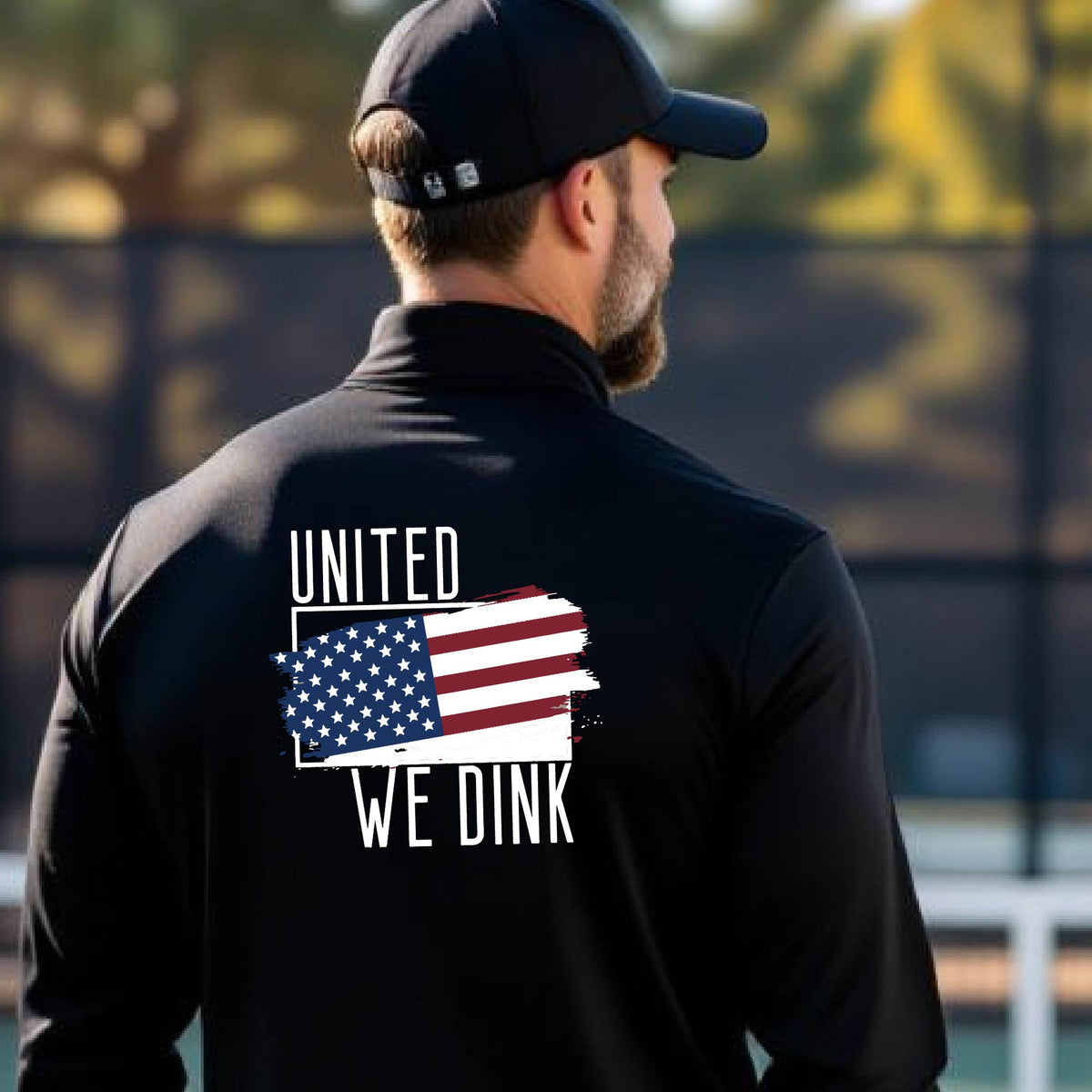 A man seen from behind on a pickleball court wearing a black quarter-zip shirt with a pickleball themed red, white and blue graphic of a patriotic flag with white words “UNITED WE DINK” above and below the flag on the back of the shirt.