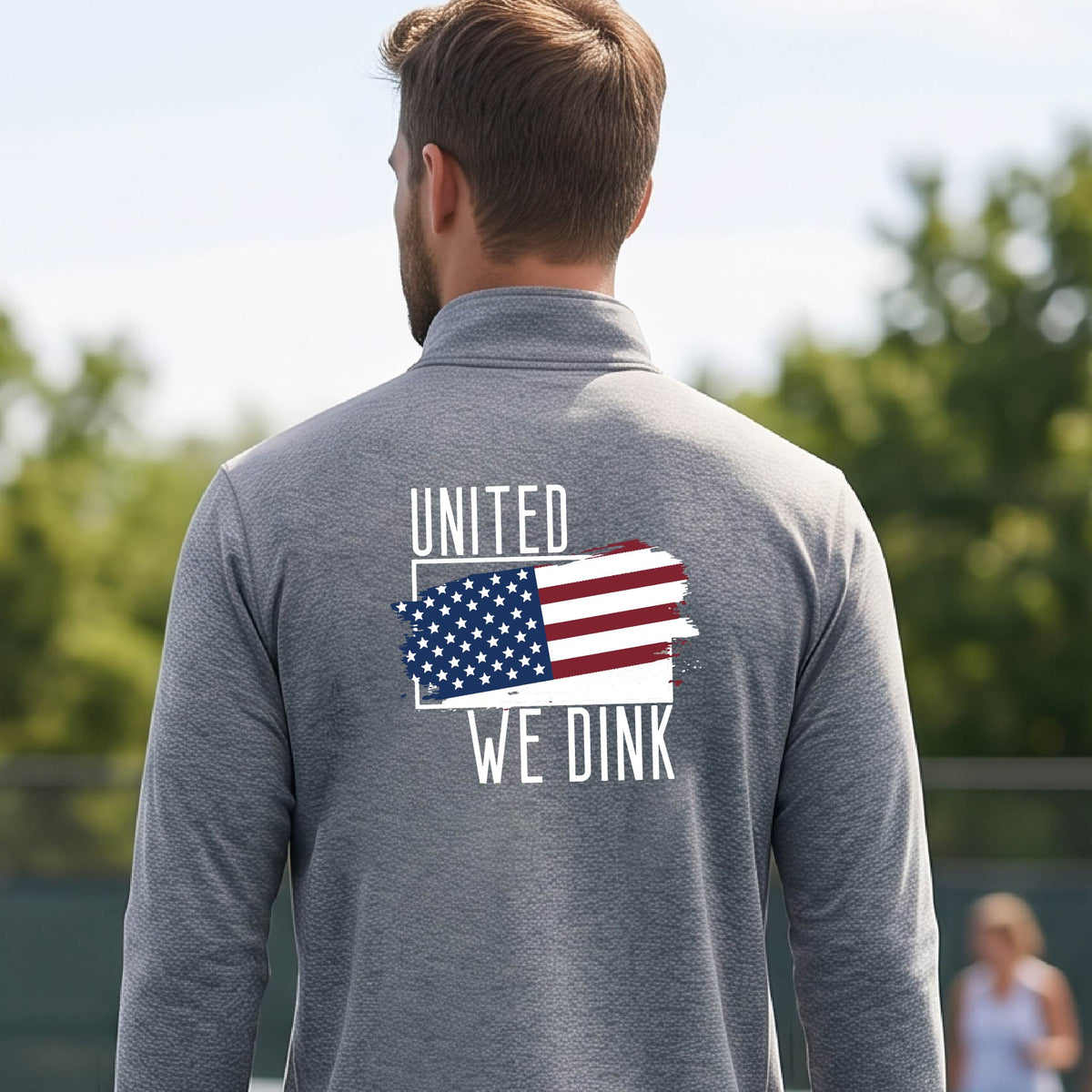 A man seen from behind on a pickleball court wearing a gray quarter-zip shirt with a pickleball themed red, white and blue graphic of a patriotic flag with white words “UNITED WE DINK” above and below the flag on the back of the shirt.