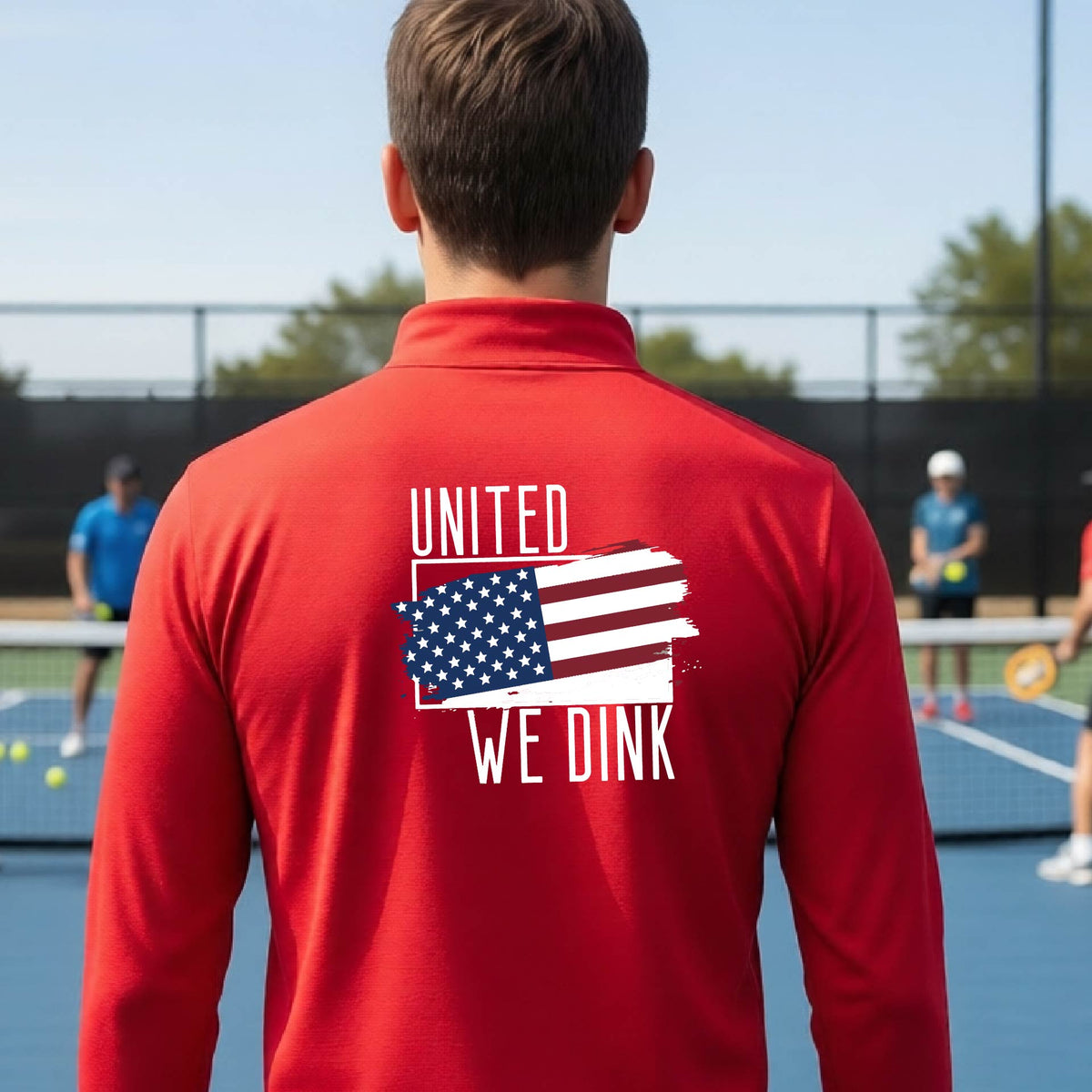 A man seen from behind on a pickleball court wearing a red quarter-zip shirt with a pickleball themed red, white and blue graphic of a patriotic flag with white words “UNITED WE DINK” above and below the flag on the back of the shirt.