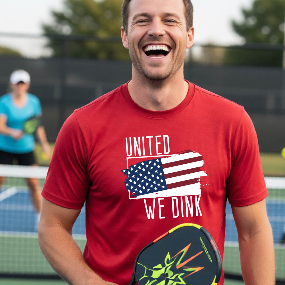 A man on a pickleball court wearing a red short-sleeve t-shirt with a pickleball themed red, white and blue graphic of a patriotic flag with white words “UNITED WE DINK” above and below the flag on the front of the shirt.