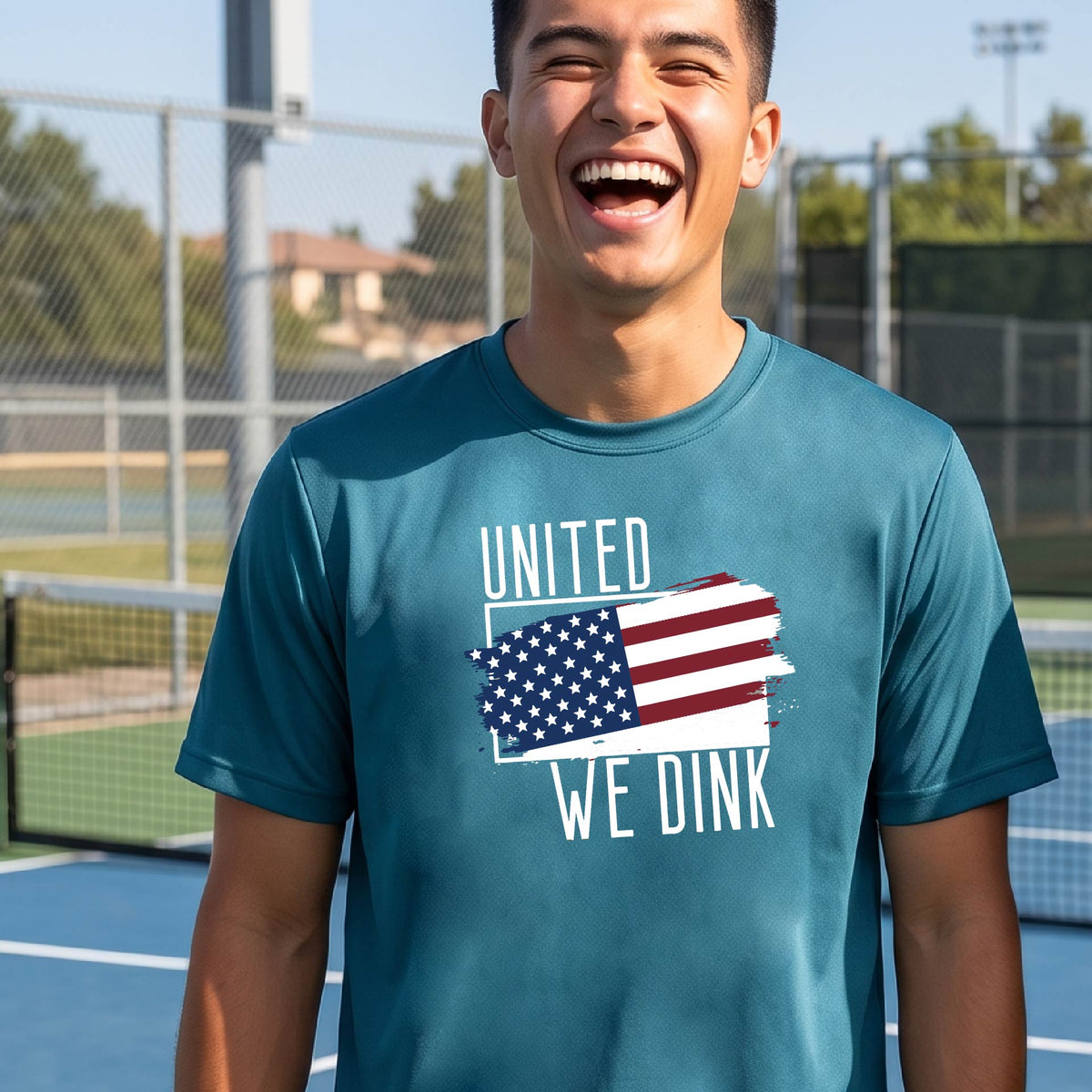 A man on a pickleball court wearing a teal short-sleeve t-shirt with a pickleball themed red, white and blue graphic of a patriotic flag with white words “UNITED WE DINK” above and below the flag on the front of the shirt.