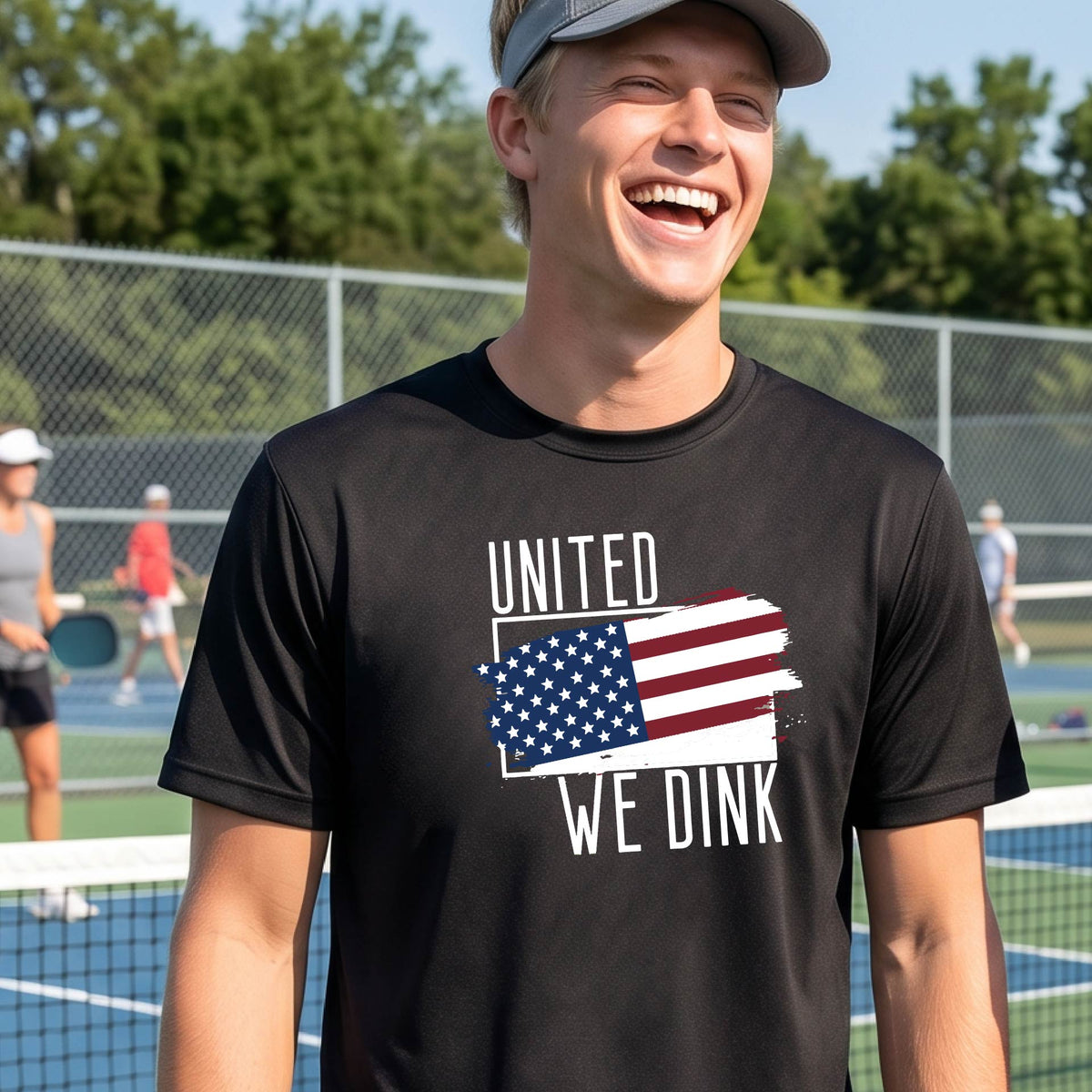 A man on a pickleball court wearing a black short-sleeve t-shirt with a pickleball themed red, white and blue graphic of a patriotic flag with white words “UNITED WE DINK” above and below the flag on the front of the shirt.
