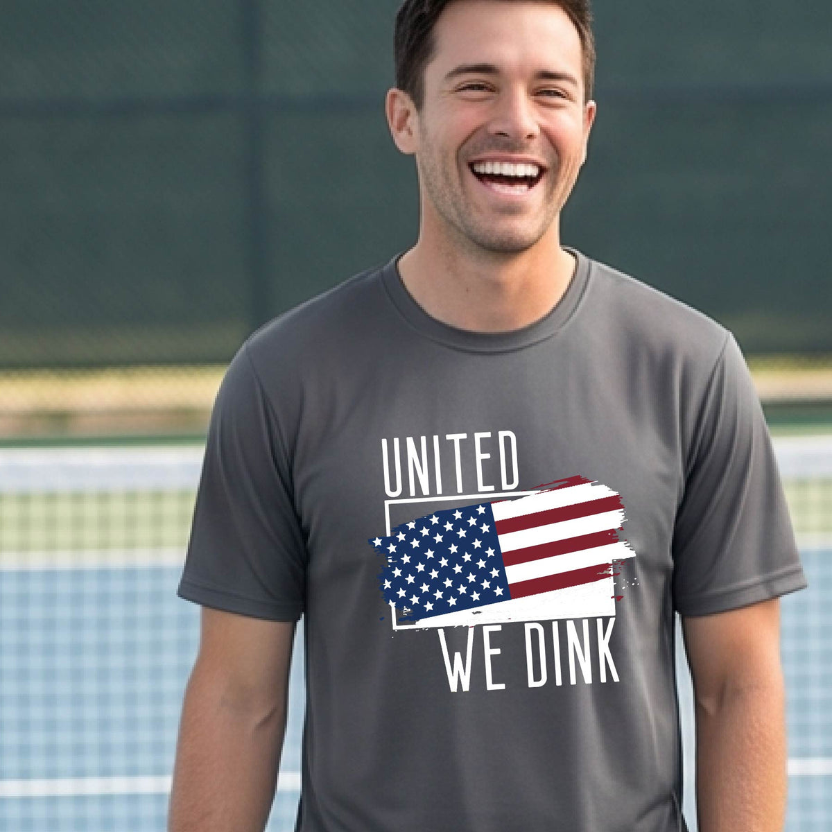 A man on a pickleball court wearing a gray short-sleeve t-shirt with a pickleball themed red, white and blue graphic of a patriotic flag with white words “UNITED WE DINK” above and below the flag on the front of the shirt.