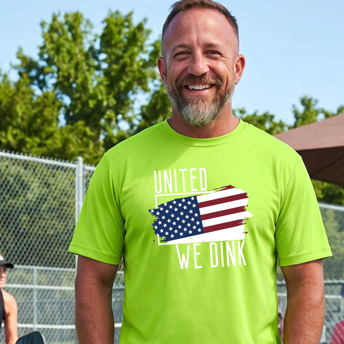 A man on a pickleball court wearing a lime green short-sleeve t-shirt with a pickleball themed red, white and blue graphic of a patriotic flag with white words “UNITED WE DINK” above and below the flag on the front of the shirt.