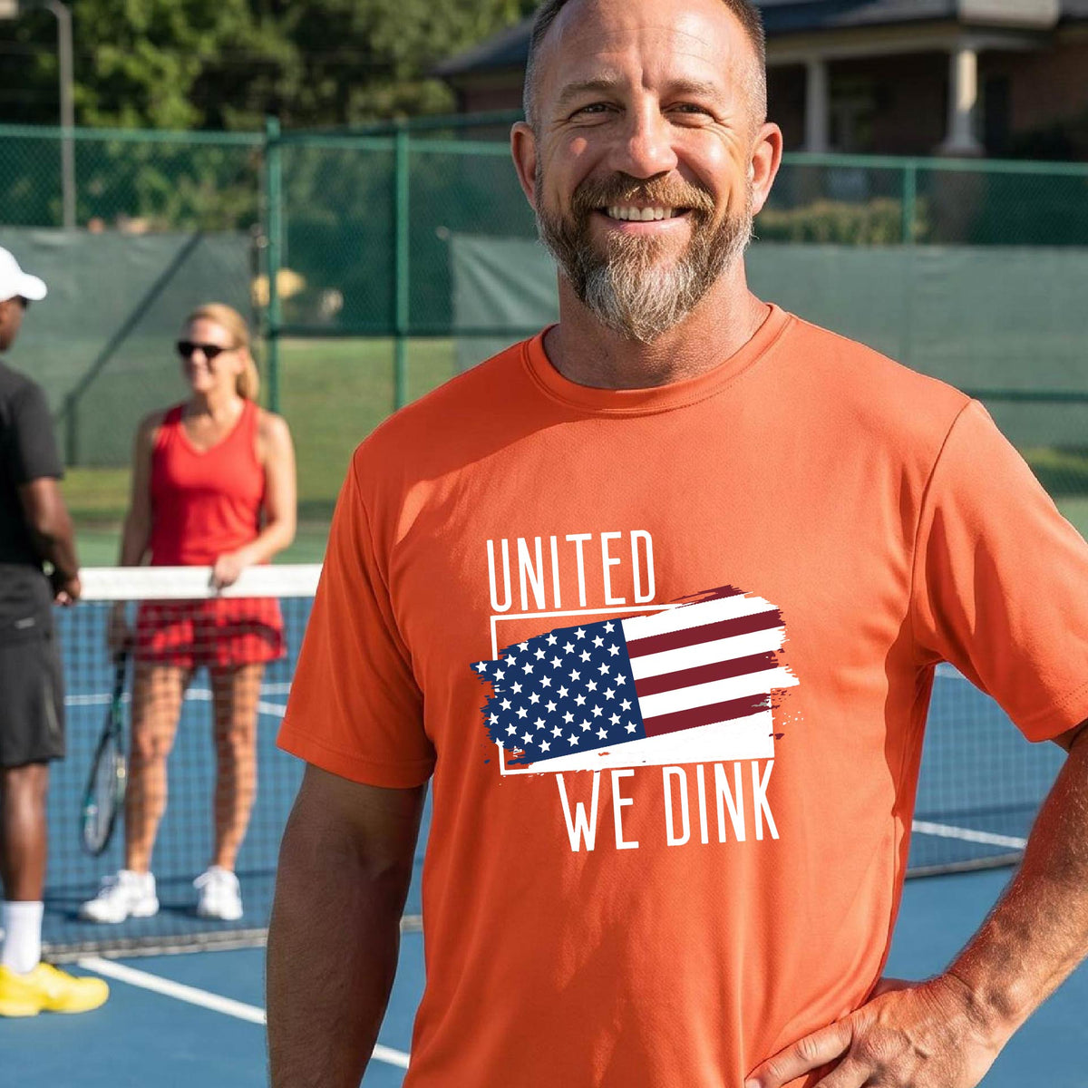 A man on a pickleball court wearing an orange short-sleeve t-shirt with a pickleball themed red, white and blue graphic of a patriotic flag with white words “UNITED WE DINK” above and below the flag on the front of the shirt.