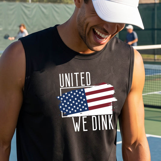 A man on a pickleball court wearing a black sleeveless t-shirt with a pickleball themed red, white and blue graphic of a patriotic flag with white words “UNITED WE DINK” above and below the flag on the front of the shirt.