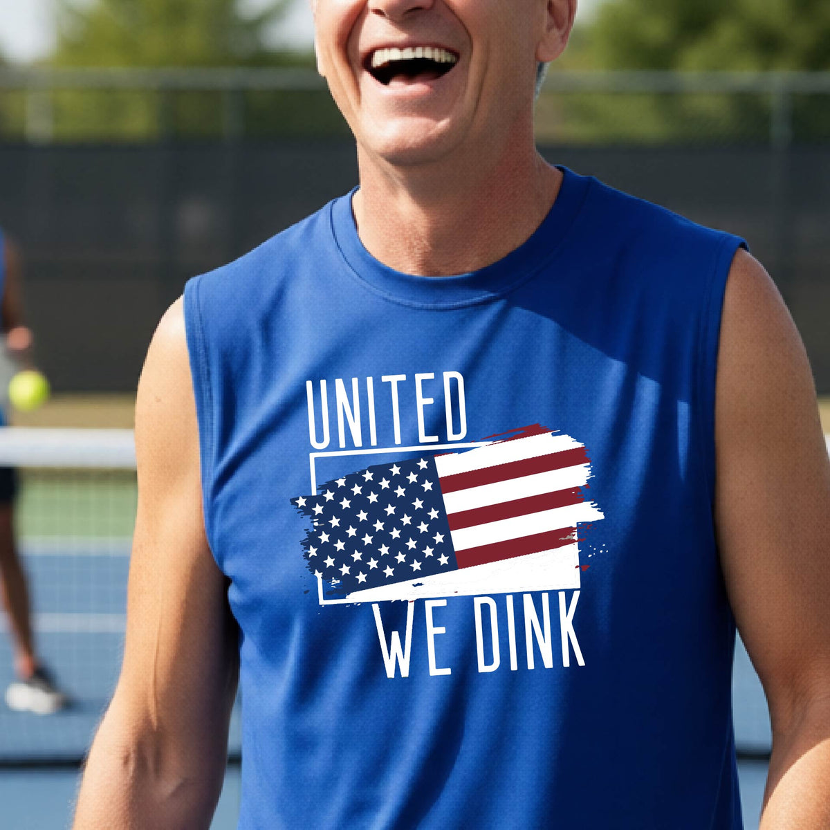A man on a pickleball court wearing a royal blue sleeveless t-shirt with a pickleball themed red, white and blue graphic of a patriotic flag with white words “UNITED WE DINK” above and below the flag on the front of the shirt.