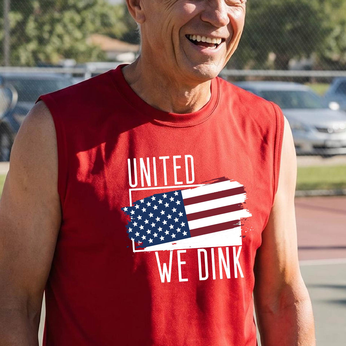 A man on a pickleball court wearing a red sleeveless t-shirt with a pickleball themed red, white and blue graphic of a patriotic flag with white words “UNITED WE DINK” above and below the flag on the front of the shirt.
