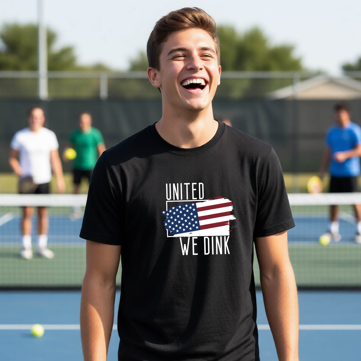 A young person on a pickleball court wearing a black short-sleeve t-shirt with a pickleball themed red, white and blue graphic of a patriotic flag with white words “UNITED WE DINK” above and below the flag, on the front of the shirt. 