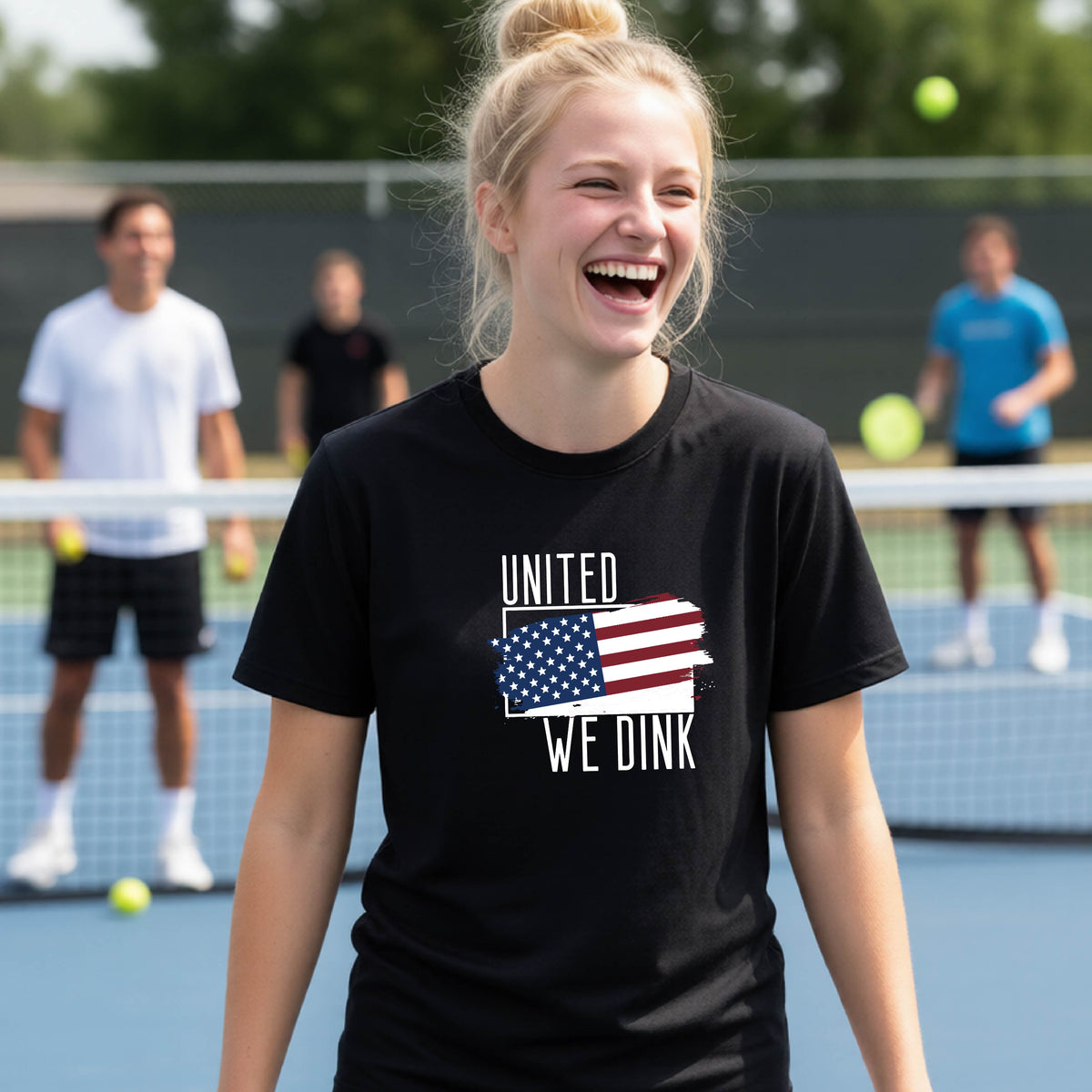 A young person on a pickleball court wearing a black short-sleeve t-shirt with a pickleball themed red, white and blue graphic of a patriotic flag with white words “UNITED WE DINK” above and below the flag, on the front of the shirt. 