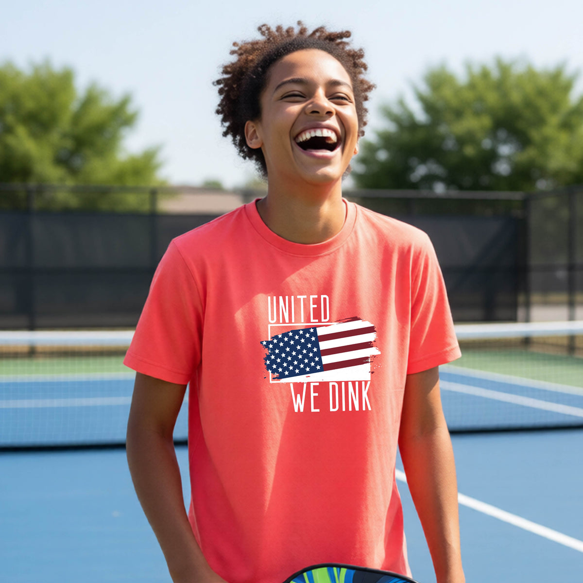 A young person on a pickleball court wearing a coral pink short-sleeve t-shirt with a pickleball themed red, white and blue graphic of a patriotic flag with white words “UNITED WE DINK” above and below the flag, on the front of the shirt. 