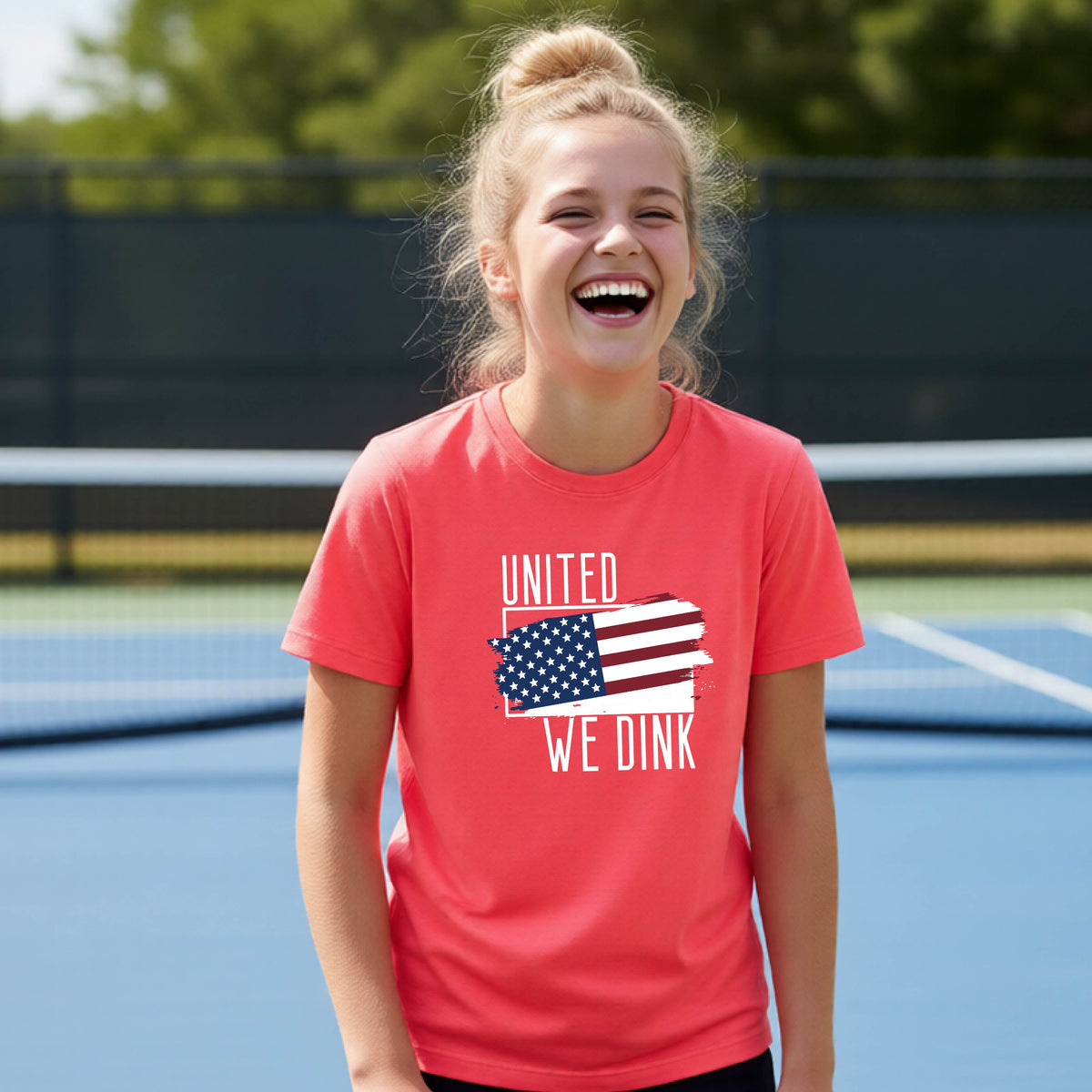 A young person on a pickleball court wearing a coral pink short-sleeve t-shirt with a pickleball themed red, white and blue graphic of a patriotic flag with white words “UNITED WE DINK” above and below the flag, on the front of the shirt. 
