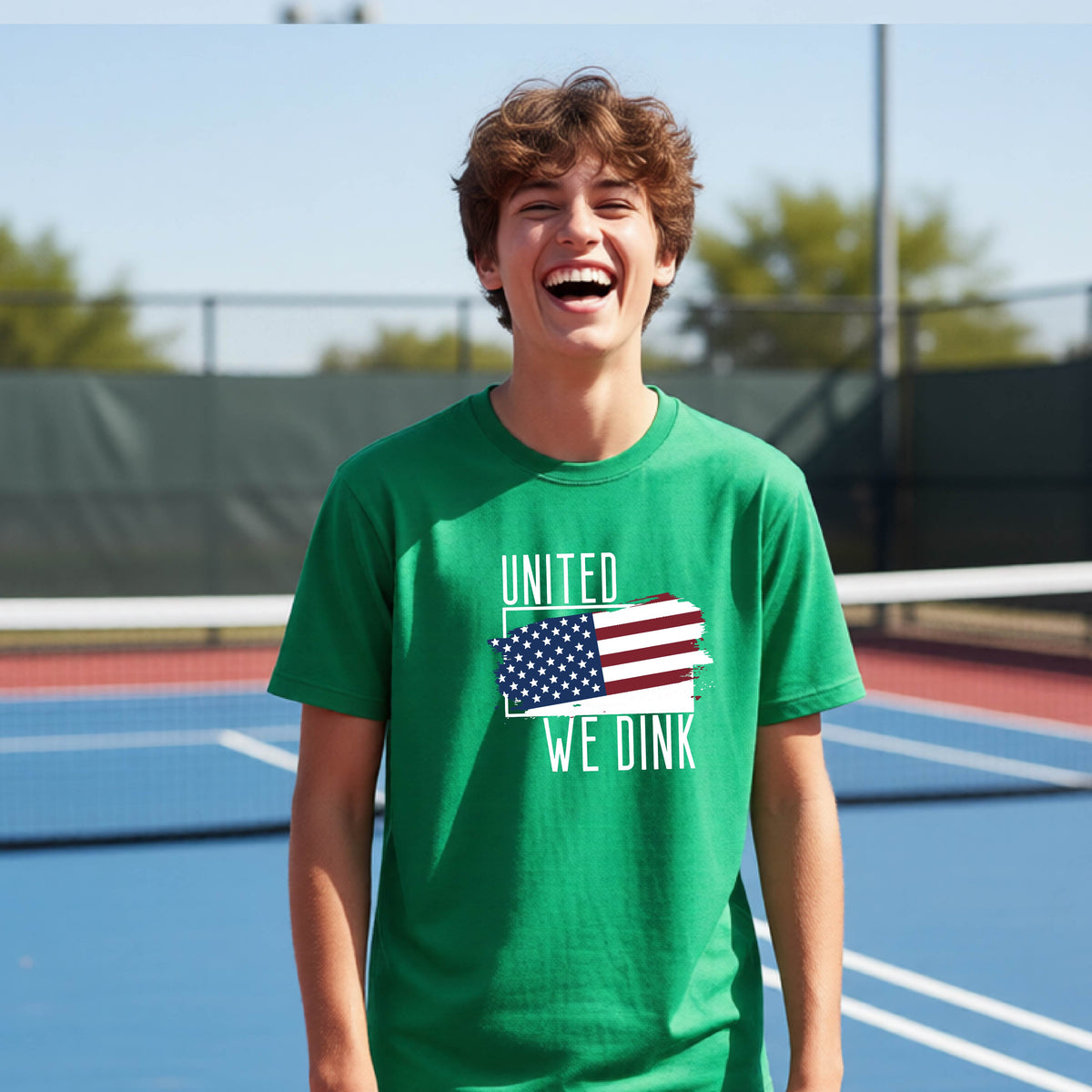 A young person on a pickleball court wearing a green short-sleeve t-shirt with a pickleball themed red, white and blue graphic of a patriotic flag with white words “UNITED WE DINK” above and below the flag, on the front of the shirt. 