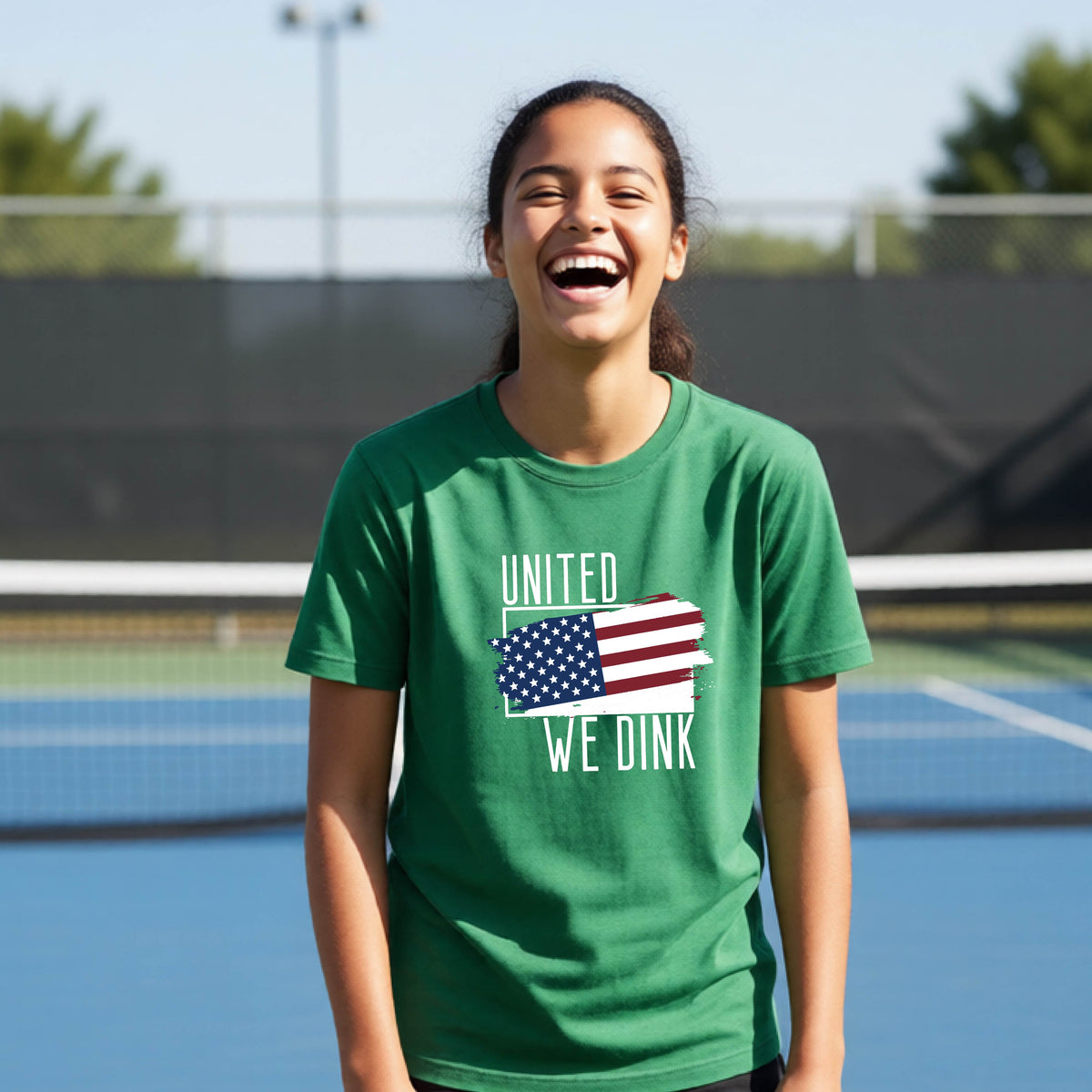 A young person on a pickleball court wearing a green short-sleeve t-shirt with a pickleball themed red, white and blue graphic of a patriotic flag with white words “UNITED WE DINK” above and below the flag, on the front of the shirt. 