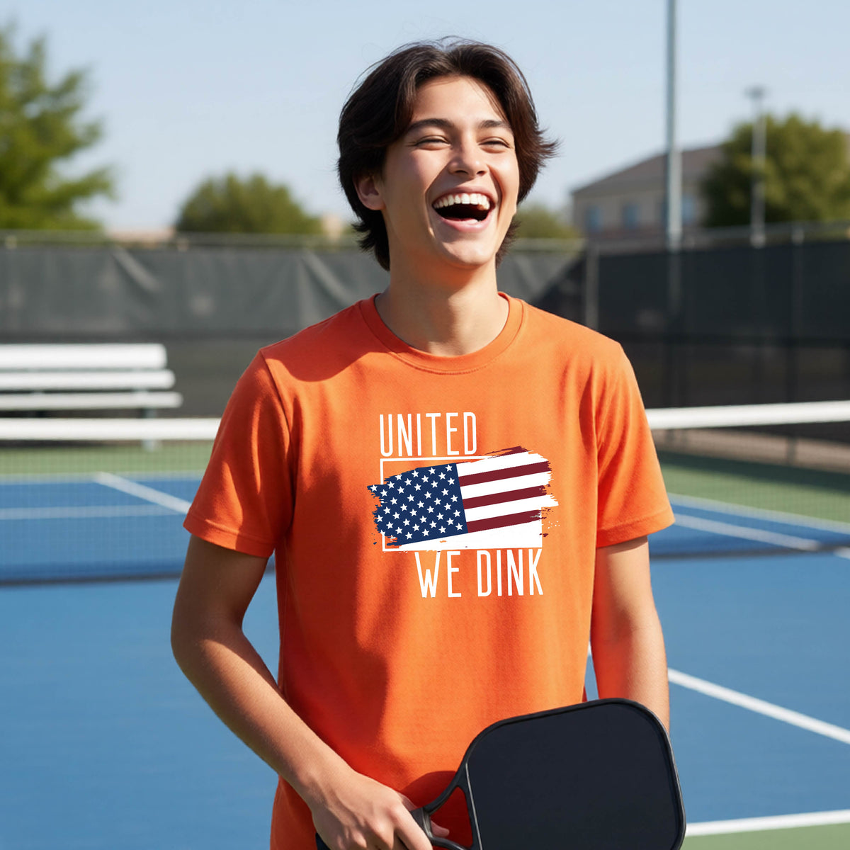 A young person on a pickleball court wearing an orange short-sleeve t-shirt with a pickleball themed red, white and blue graphic of a patriotic flag with white words “UNITED WE DINK” above and below the flag, on the front of the shirt. 