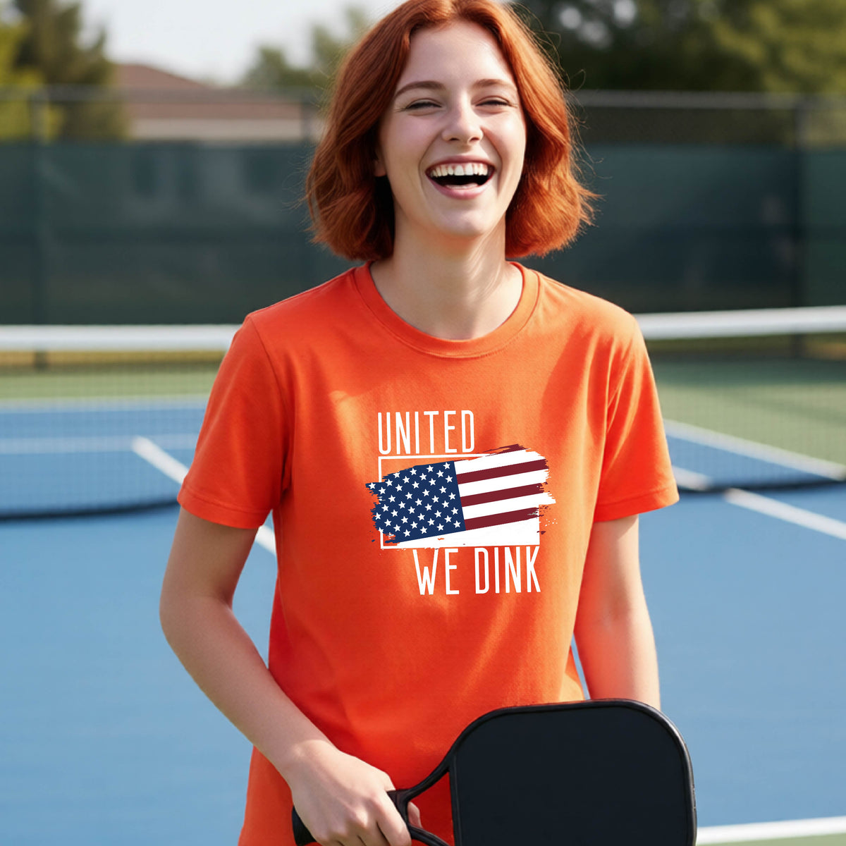 A young person on a pickleball court wearing an orange short-sleeve t-shirt with a pickleball themed red, white and blue graphic of a patriotic flag with white words “UNITED WE DINK” above and below the flag, on the front of the shirt. 