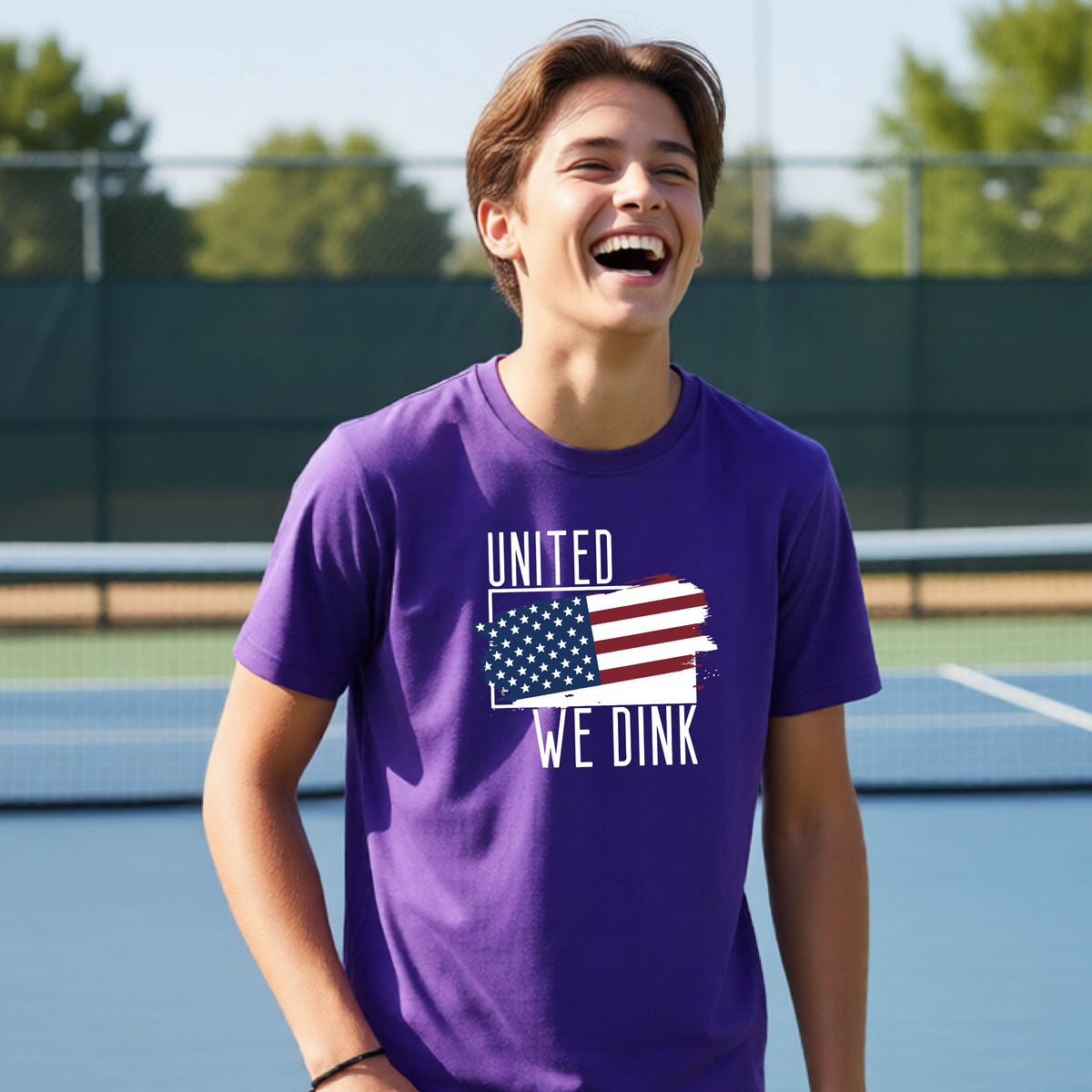 A young person on a pickleball court wearing a purple short-sleeve t-shirt with a pickleball themed red, white and blue graphic of a patriotic flag with white words “UNITED WE DINK” above and below the flag, on the front of the shirt. 