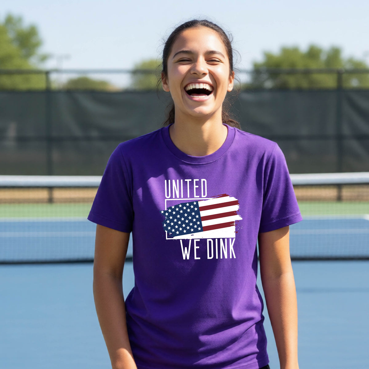 A young person on a pickleball court wearing a purple short-sleeve t-shirt with a pickleball themed red, white and blue graphic of a patriotic flag with white words “UNITED WE DINK” above and below the flag, on the front of the shirt. 
