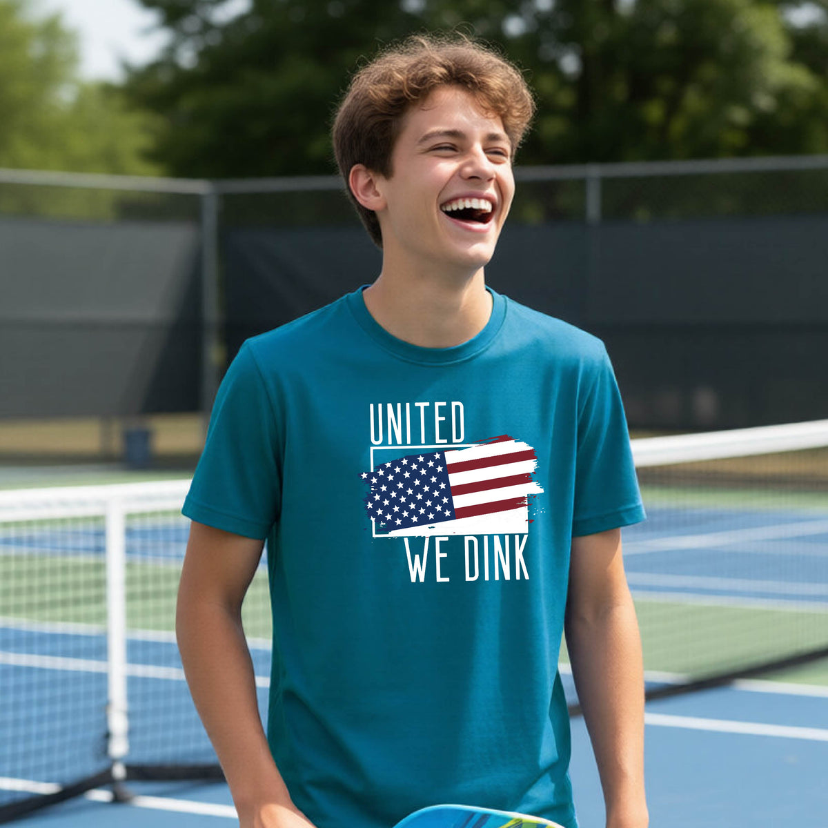A young person on a pickleball court wearing a teal short-sleeve t-shirt with a pickleball themed red, white and blue graphic of a patriotic flag with white words “UNITED WE DINK” above and below the flag, on the front of the shirt. 