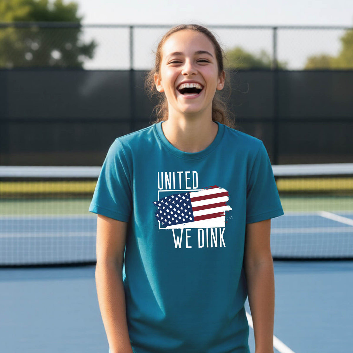 A young person on a pickleball court wearing a teal short-sleeve t-shirt with a pickleball themed red, white and blue graphic of a patriotic flag with white words “UNITED WE DINK” above and below the flag, on the front of the shirt. 