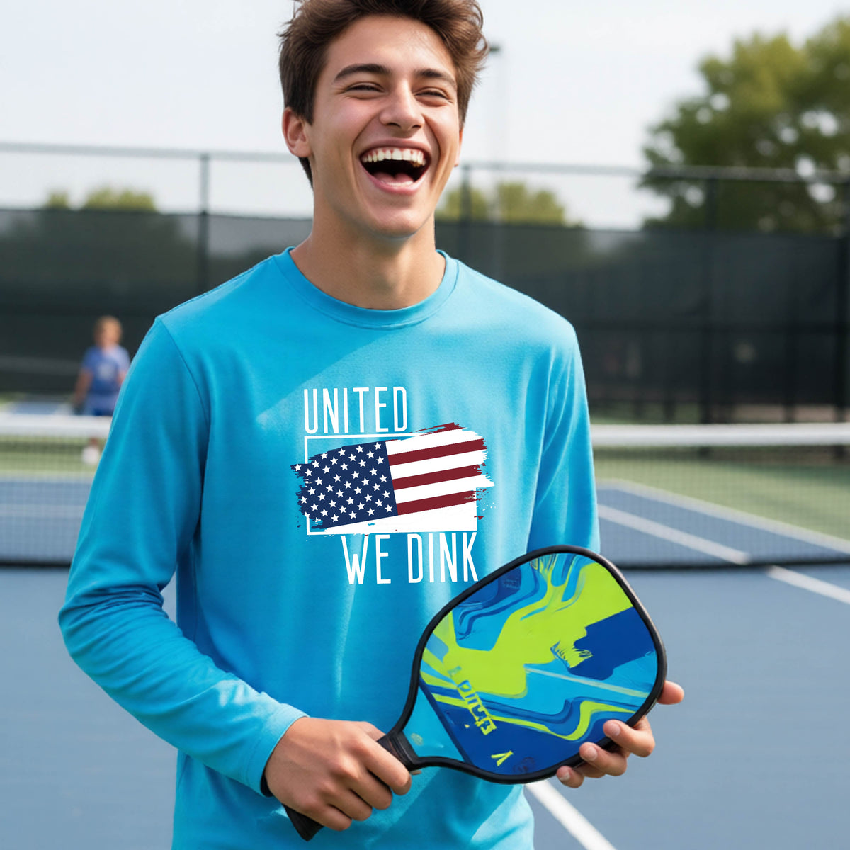 A young person on a pickleball court wearing a bright blue long-sleeve t-shirt with a pickleball themed red, white and blue graphic of a patriotic flag with white words “UNITED WE DINK” above and below the flag, on the front of the shirt. 