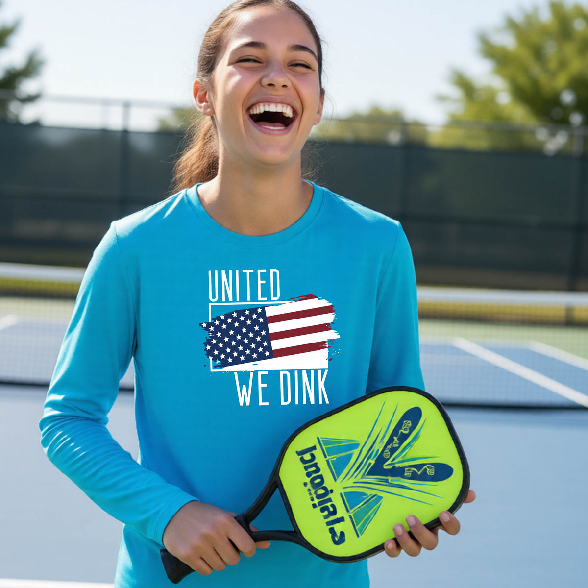 A young person on a pickleball court wearing a bright blue  long-sleeve t-shirt with a pickleball themed red, white and blue graphic of a patriotic flag with white words “UNITED WE DINK” above and below the flag, on the front of the shirt. 