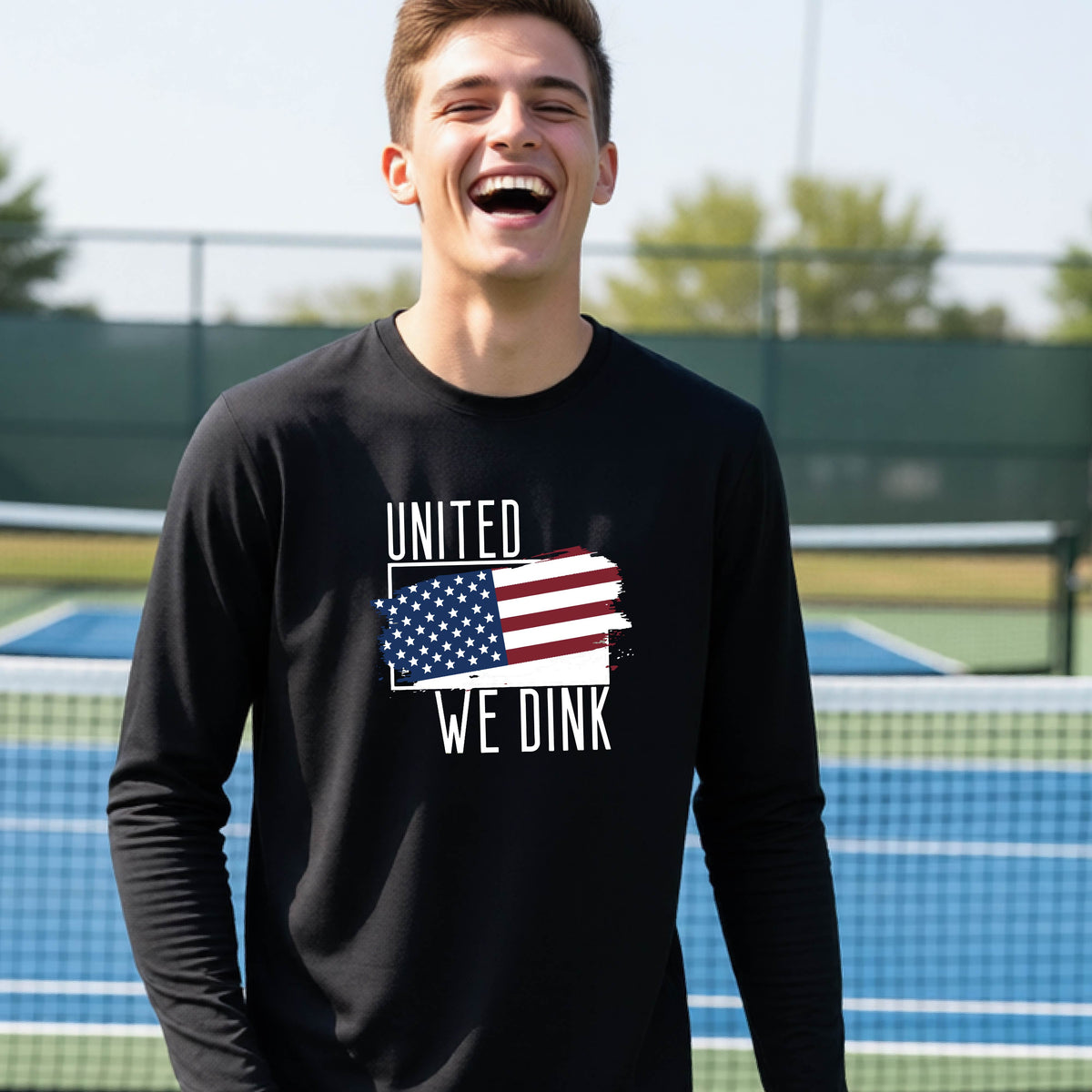 A young person on a pickleball court wearing a black long-sleeve t-shirt with a pickleball themed red, white and blue graphic of a patriotic flag with white words “UNITED WE DINK” above and below the flag, on the front of the shirt. 