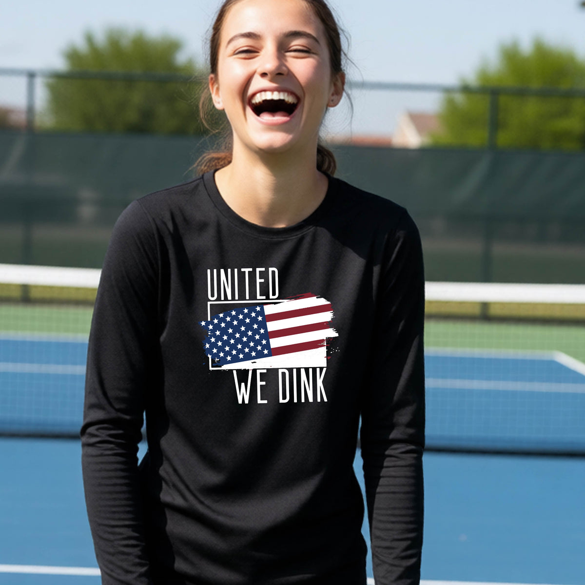 A young person on a pickleball court wearing a black long-sleeve t-shirt with a pickleball themed red, white and blue graphic of a patriotic flag with white words “UNITED WE DINK” above and below the flag, on the front of the shirt. 