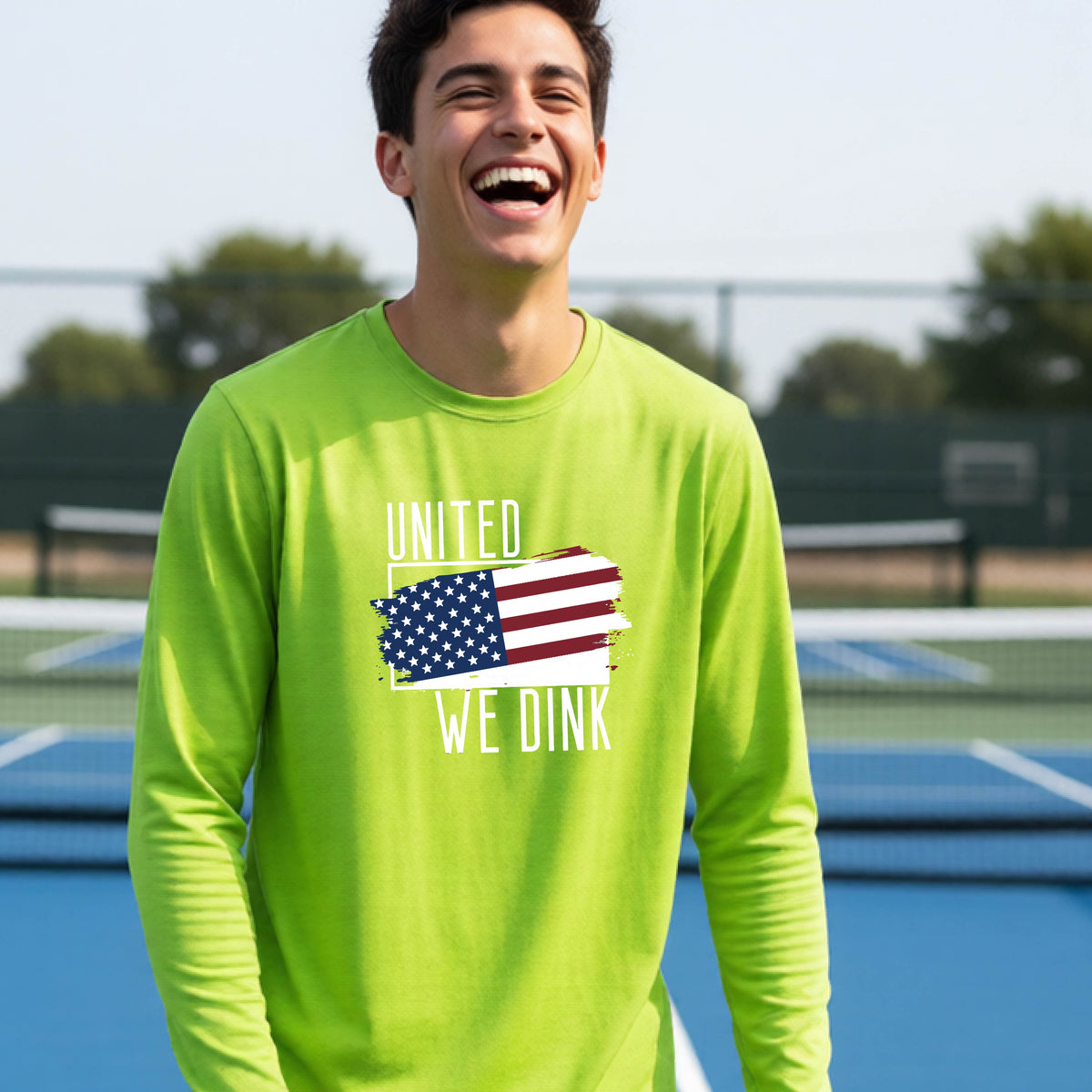 A young person on a pickleball court wearing a lime green long-sleeve t-shirt with a pickleball themed red, white and blue graphic of a patriotic flag with white words “UNITED WE DINK” above and below the flag, on the front of the shirt. 