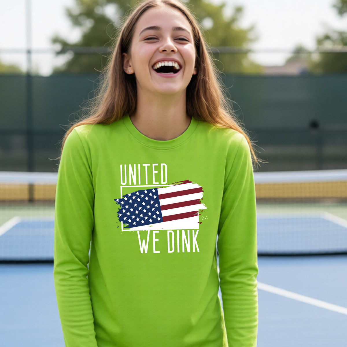 A young person on a pickleball court wearing a lime green long-sleeve t-shirt with a pickleball themed red, white and blue graphic of a patriotic flag with white words “UNITED WE DINK” above and below the flag, on the front of the shirt. 