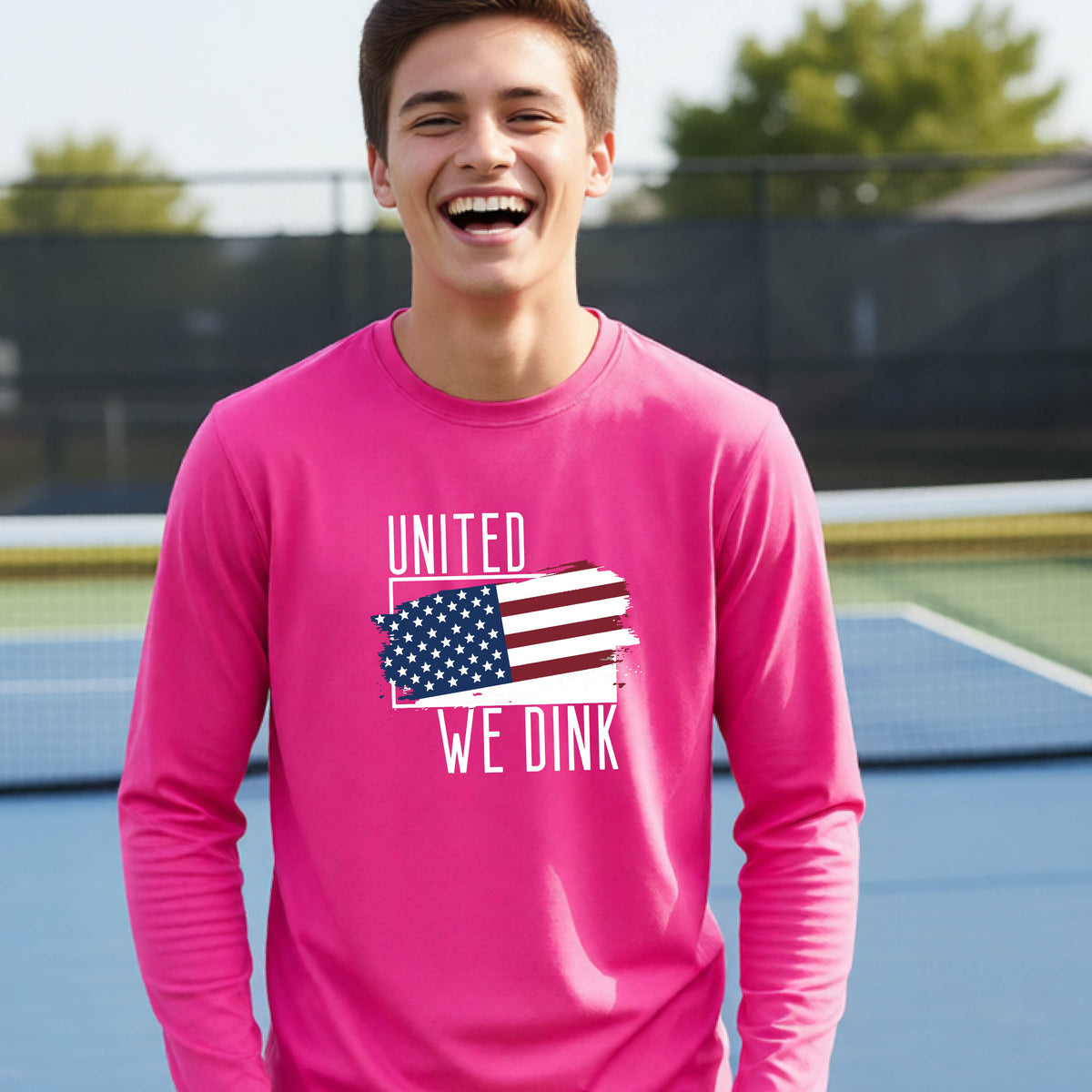 A young person on a pickleball court wearing a pink long-sleeve t-shirt with a pickleball themed red, white and blue graphic of a patriotic flag with white words “UNITED WE DINK” above and below the flag, on the front of the shirt. 