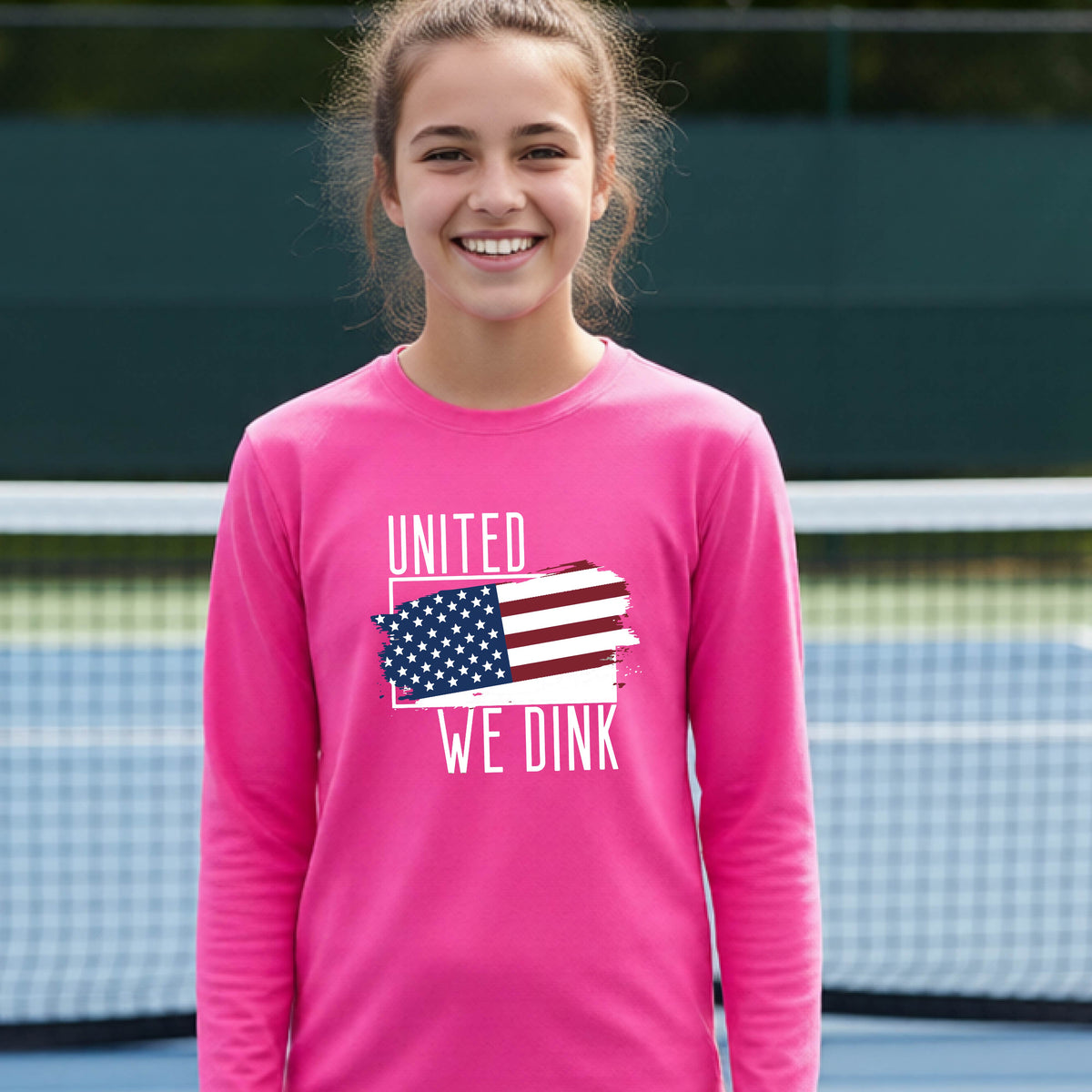 A young person on a pickleball court wearing a pink long-sleeve t-shirt with a pickleball themed red, white and blue graphic of a patriotic flag with white words “UNITED WE DINK” above and below the flag, on the front of the shirt. 