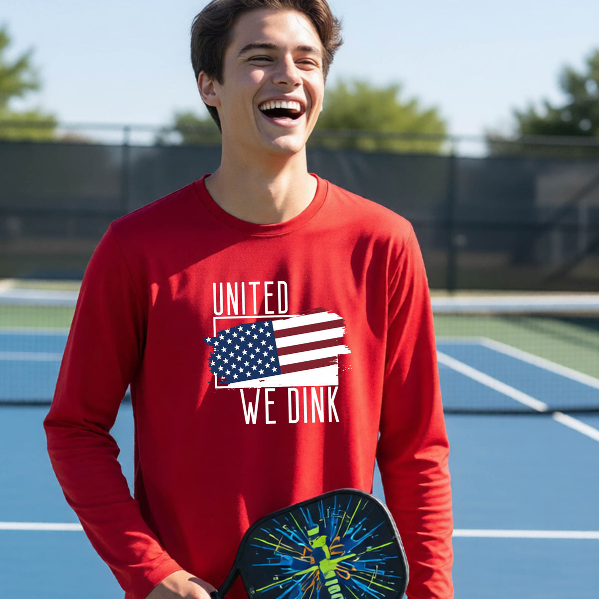 A young person on a pickleball court wearing a red long-sleeve t-shirt with a pickleball themed red, white and blue graphic of a patriotic flag with white words “UNITED WE DINK” above and below the flag, on the front of the shirt. 


