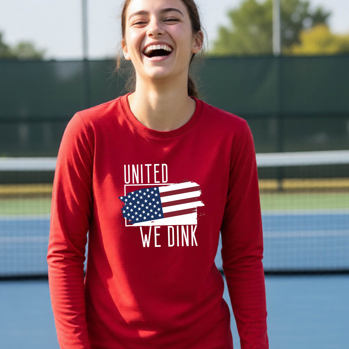 A young person on a pickleball court wearing a red long-sleeve t-shirt with a pickleball themed red, white and blue graphic of a patriotic flag with white words “UNITED WE DINK” above and below the flag, on the front of the shirt. 