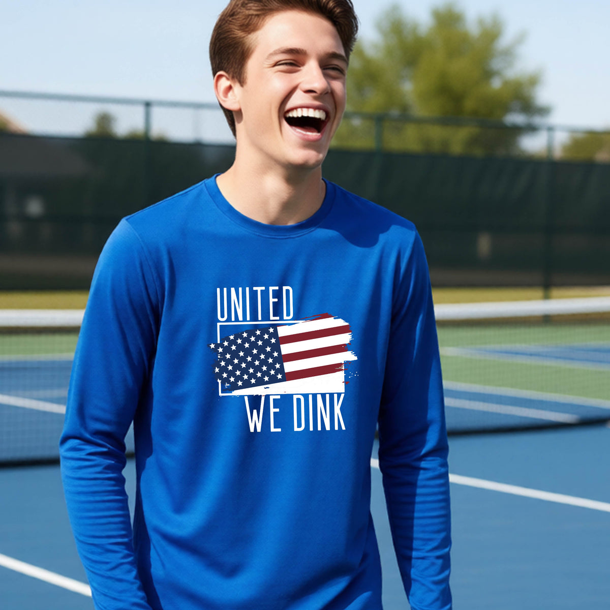 A young person on a pickleball court wearing a royal blue long-sleeve t-shirt with a pickleball themed red, white and blue graphic of a patriotic flag with white words “UNITED WE DINK” above and below the flag, on the front of the shirt. 