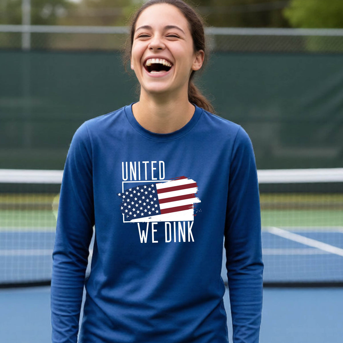 A young person on a pickleball court wearing a royal blue long-sleeve t-shirt with a pickleball themed red, white and blue graphic of a patriotic flag with white words “UNITED WE DINK” above and below the flag, on the front of the shirt. 
