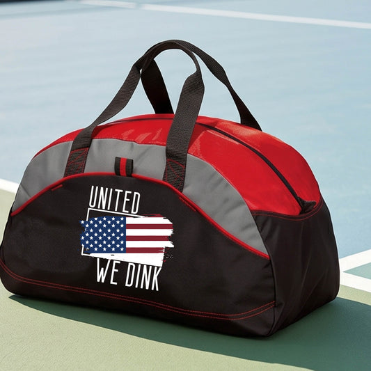A close-up of a medium size red and black tote bag with gray panels with a pickleball themed red, white and blue graphic of a patriotic flag with white words “UNITED WE DINK” above and below the flag on the front of the bag.

